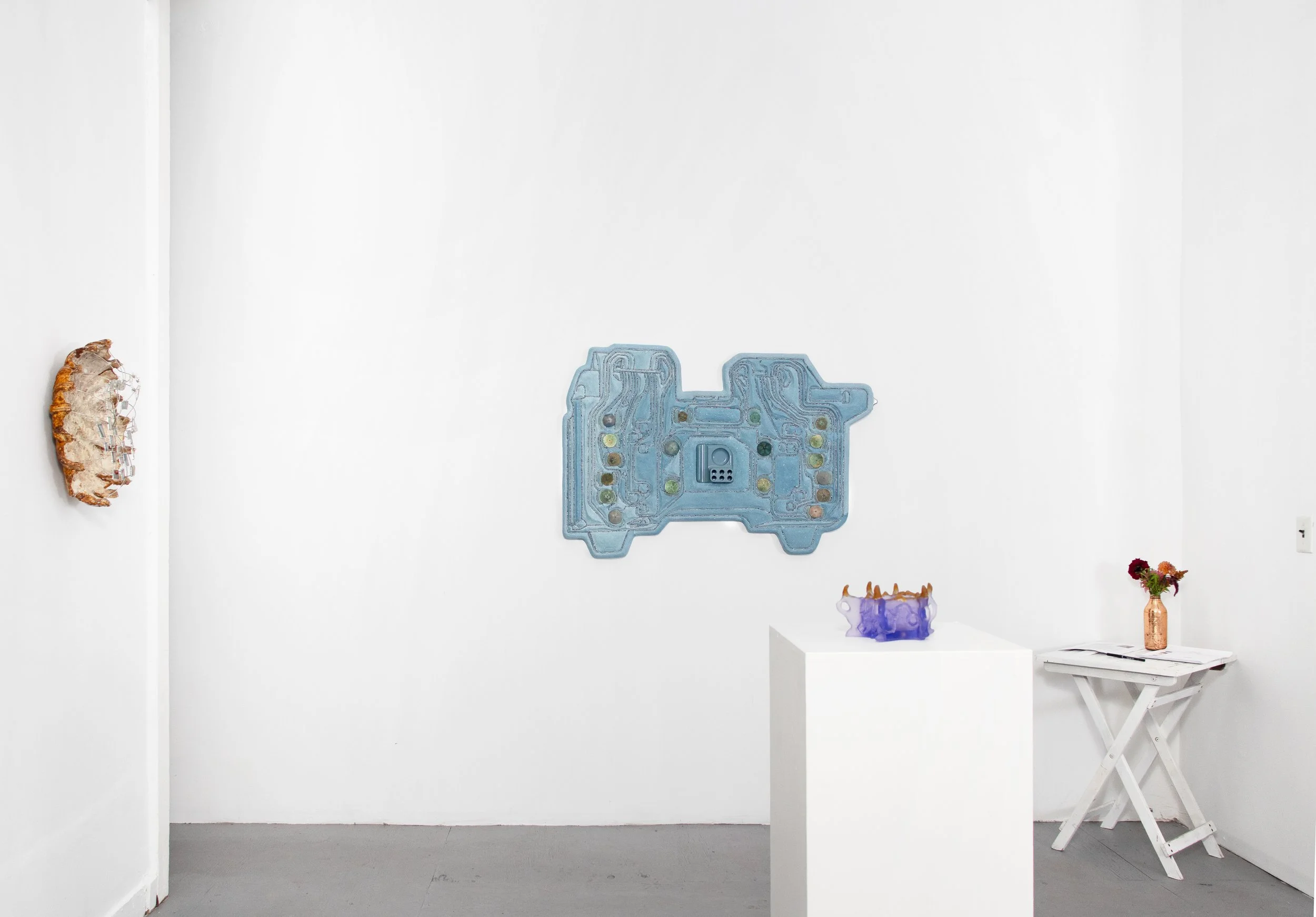 Art gallery with a white wall displaying abstract sculptures, including a wall-mounted piece, a purple glass sculpture on a white pedestal, and a small orange vase with red and purple flowers on a white table.