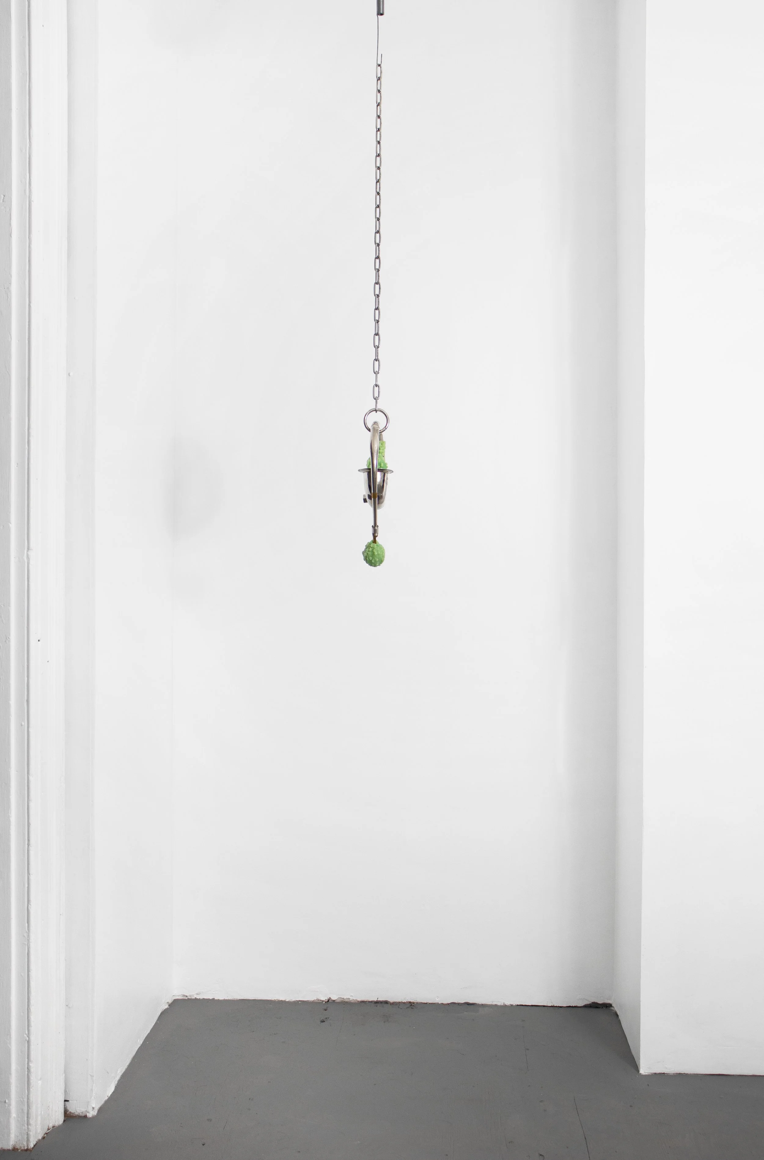 A green ball attached to a metal hook hanging from a chain in a white-walled room.