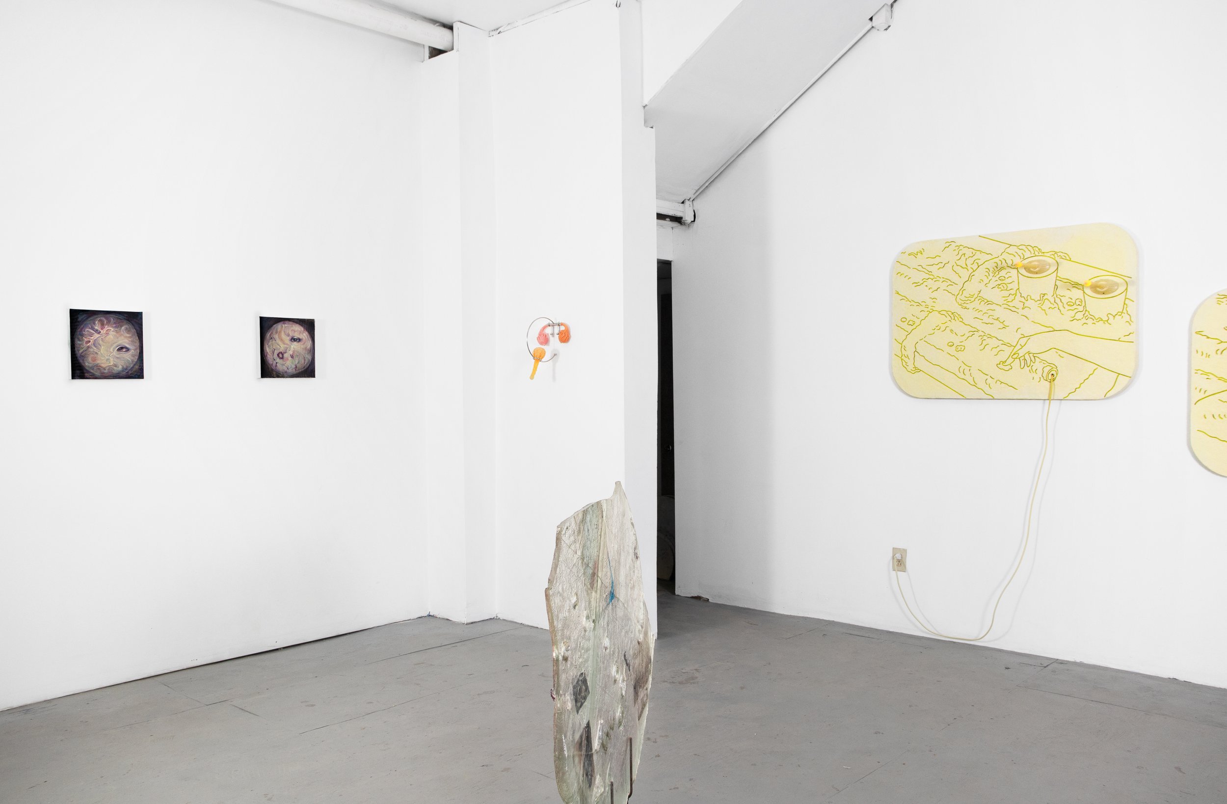 An art gallery with white walls and concrete floors. There are two framed artworks on the left wall, a small sculpture in the center of the room, and several line drawings with yellow background on the right wall. The drawings include a hand and seve