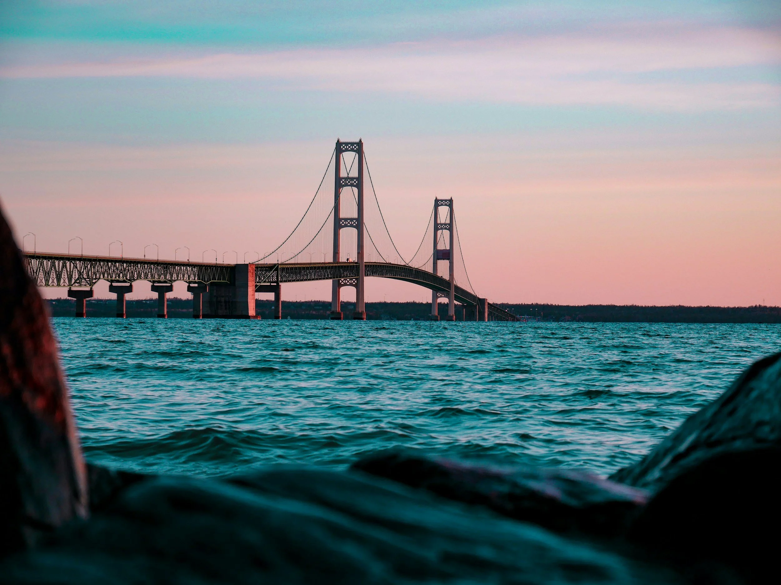 A view of a large suspension bridge over a body of water at sunset, with the sky painted in pink and blue hues.