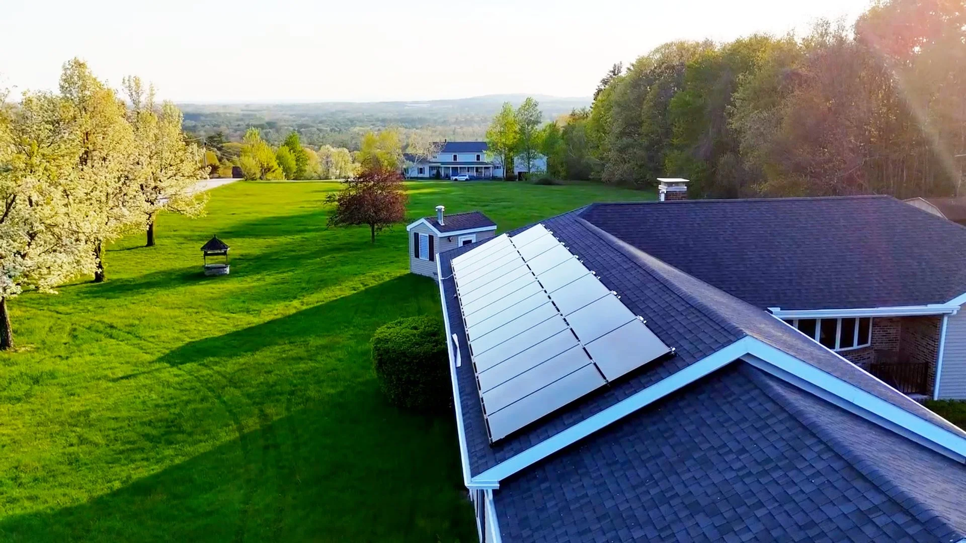 A house with solar panels on its roof, surrounded by green grass, trees, and a lawn ornament, in a rural landscape with other houses in the distance.
