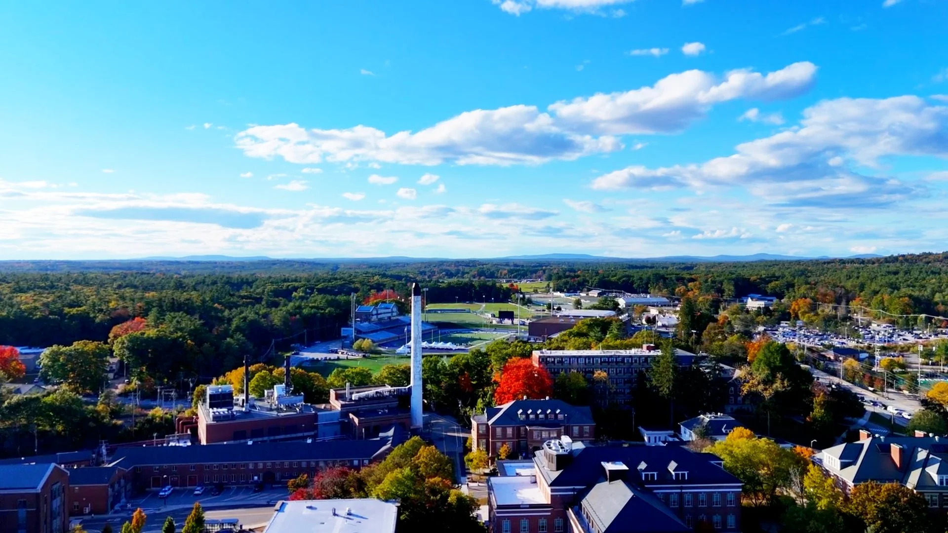 Aerial view of a cityscape with green trees, buildings, parking lots, a stadium, and distant rolling hills under a partly cloudy sky.