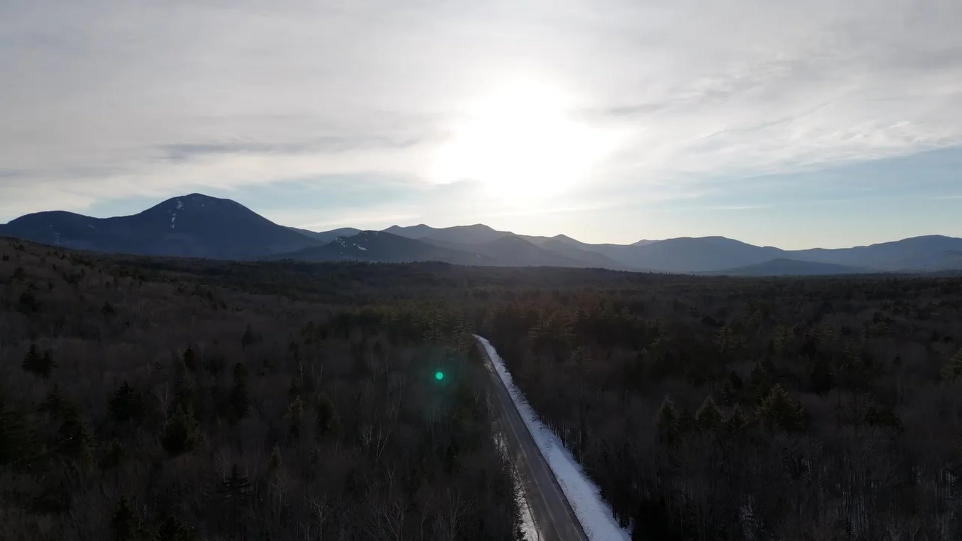 Aerial view of a mountain landscape with snow-capped peaks, a forested area, and a long straight road running through the trees, with the sun shining brightly in the sky.