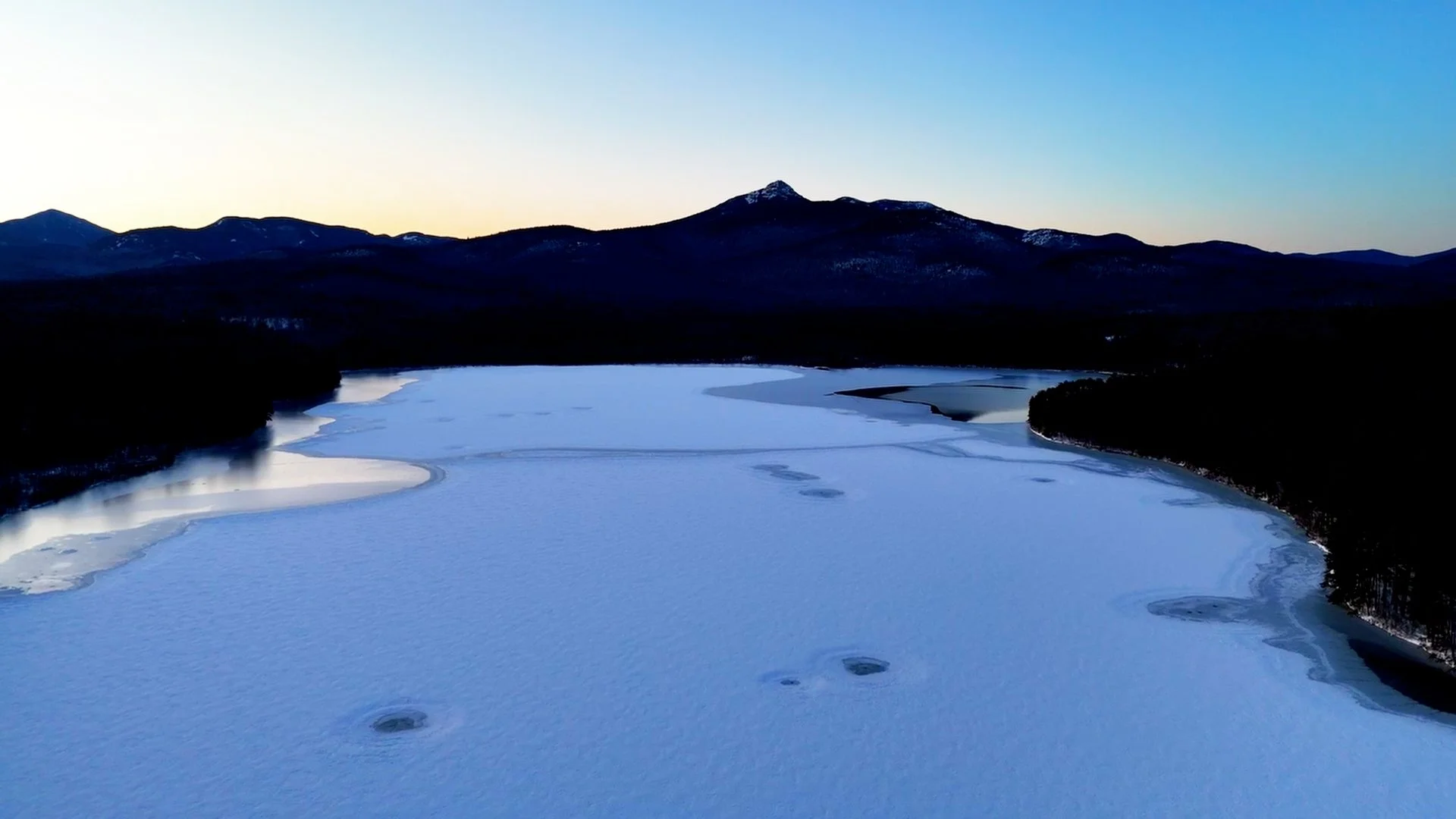 A frozen lake surrounded by snow-covered land, with mountains in the background under a clear sky at dusk.