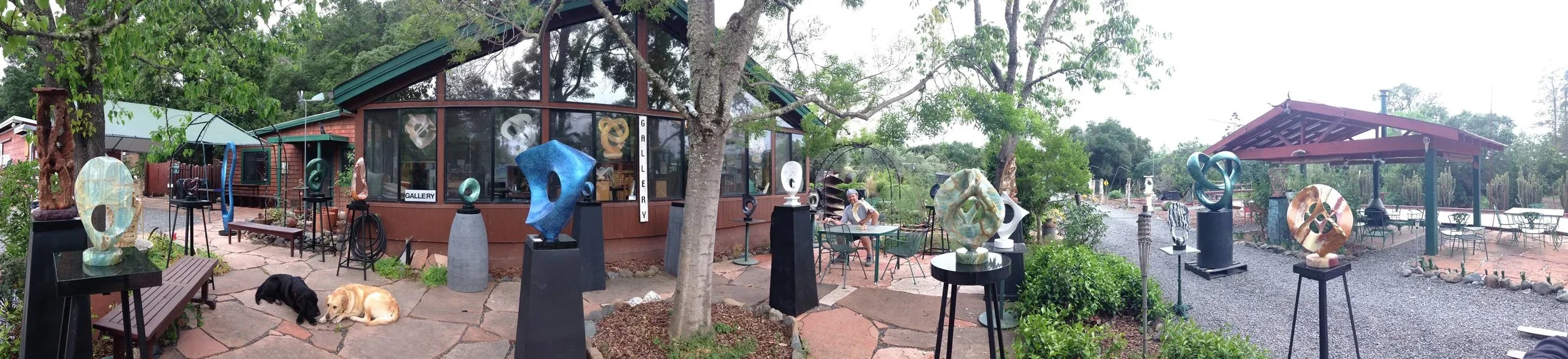 Outdoor art gallery with sculptures displayed on pedestals and tables, surrounded by trees and benches. A person sits at a table, and two dogs are resting on the ground.