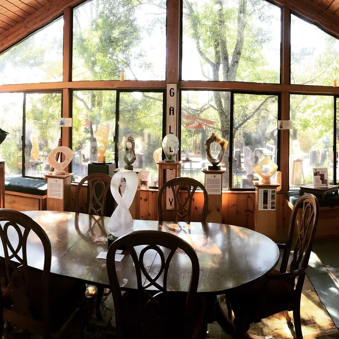 Sunlit room with large windows displaying trees outside, a wooden dining table with six chairs, and various abstract sculptures on display stands along the windows.