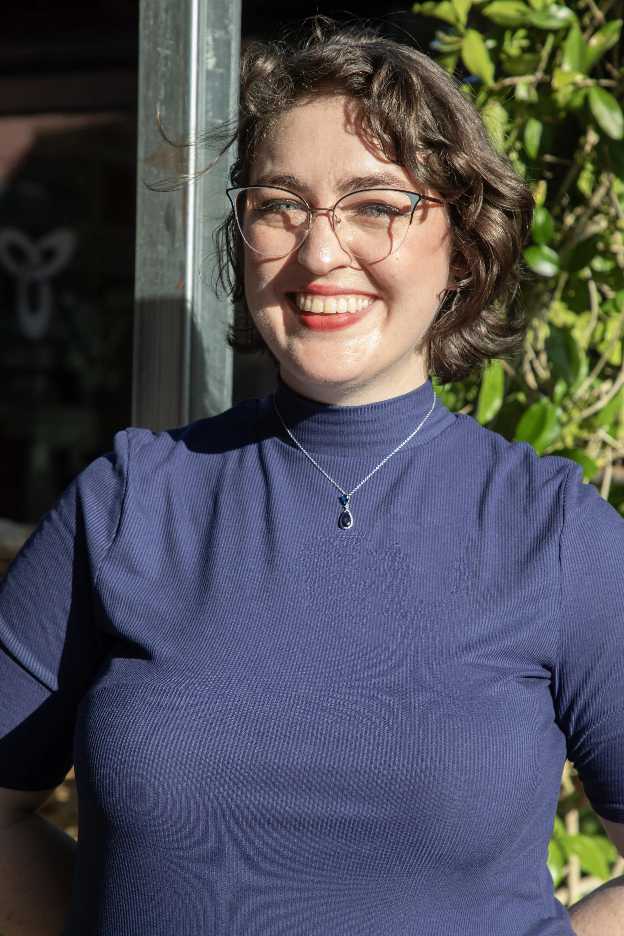 A smiling woman with glasses, short curly brown hair, wearing a blue turtleneck and a necklace with a teardrop pendant, standing outdoors with greenery and sunlight.