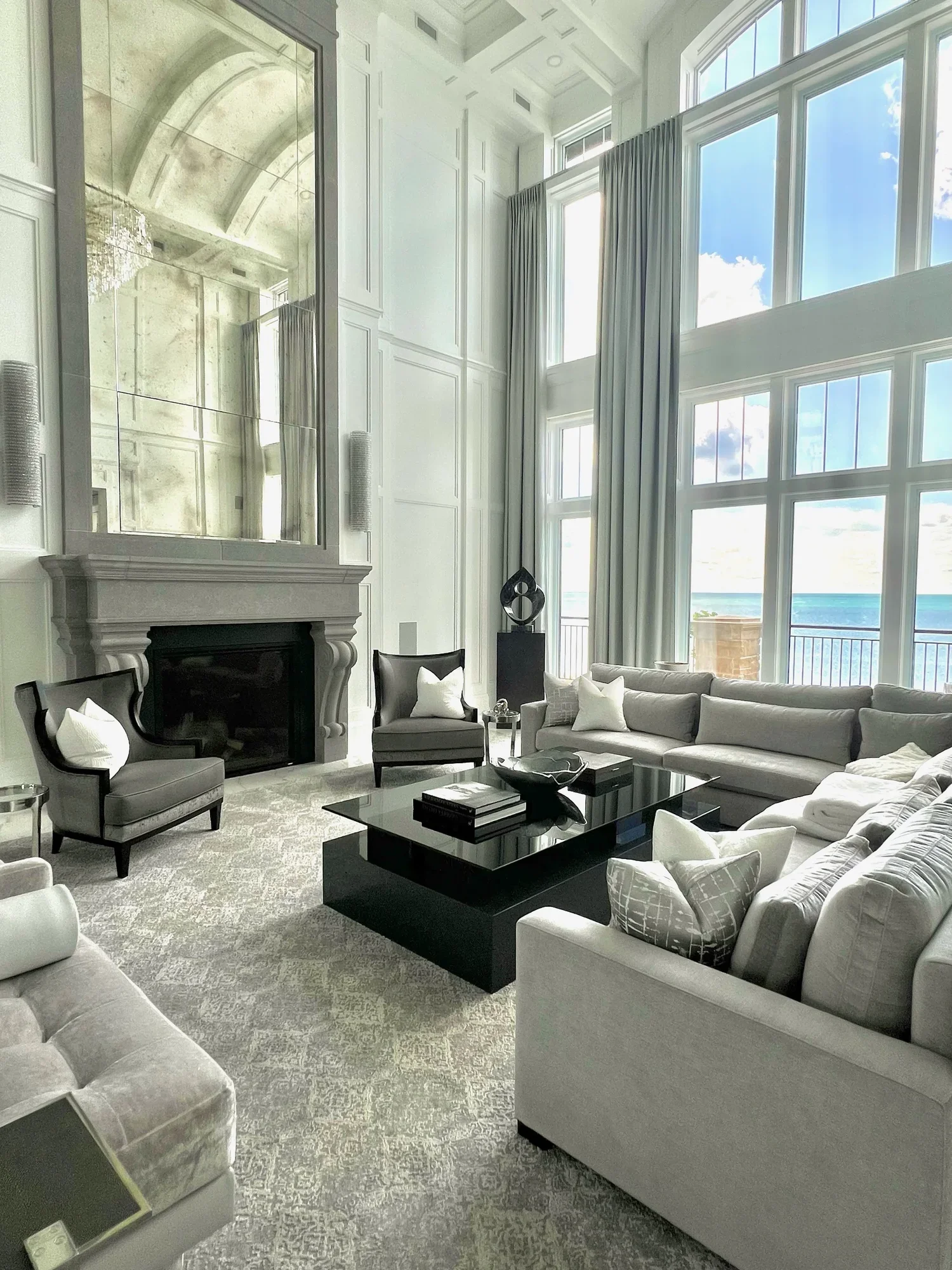 Bright living room with large windows revealing an ocean view, high ceiling, white walls, fireplace with mirror above, and modern gray sofas and chairs with white pillows.