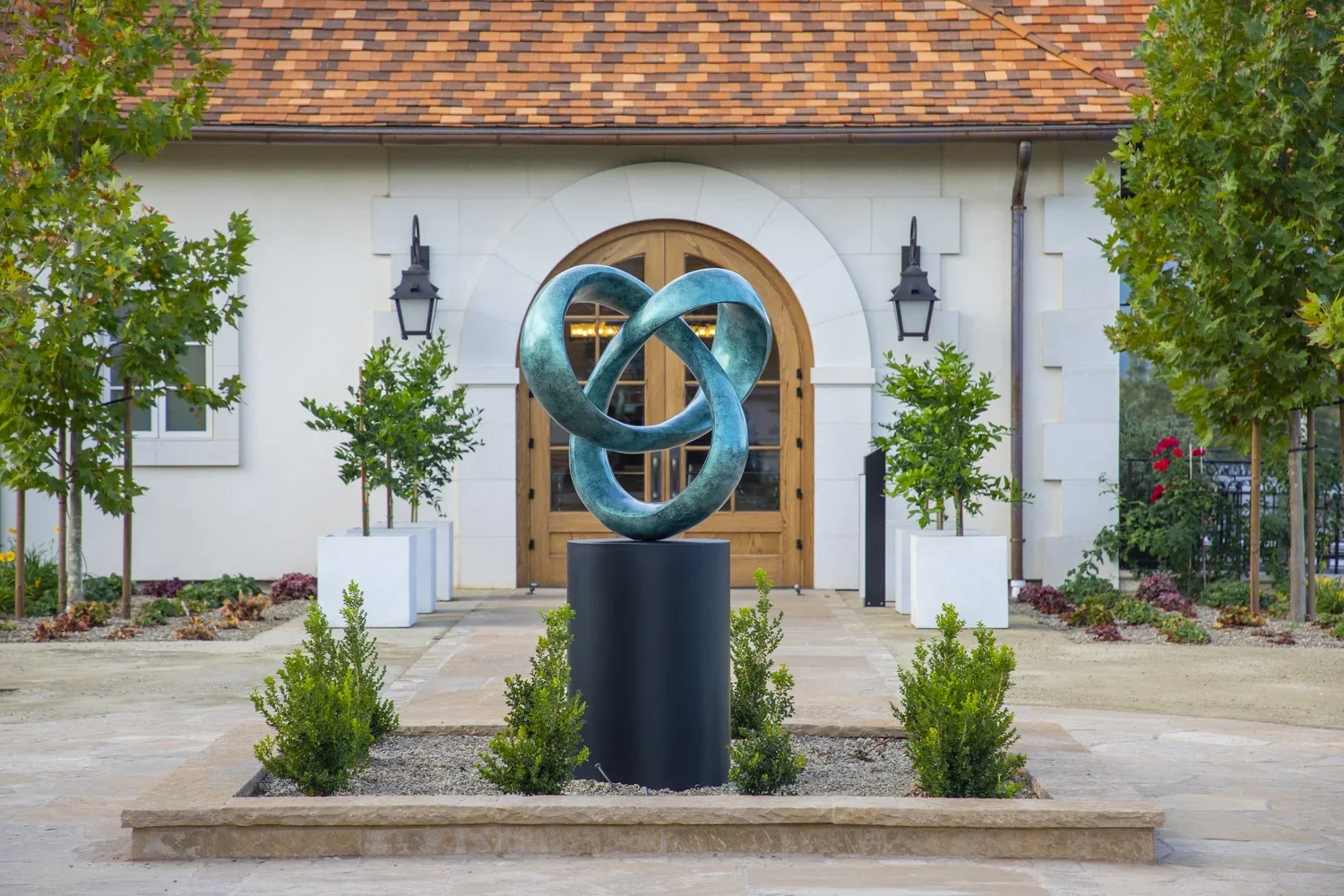 A modern outdoor sculpture of interlocked, twisted loops made of metal, situated on a black pedestal in front of a house with a wooden door, surrounded by trees and potted plants.
