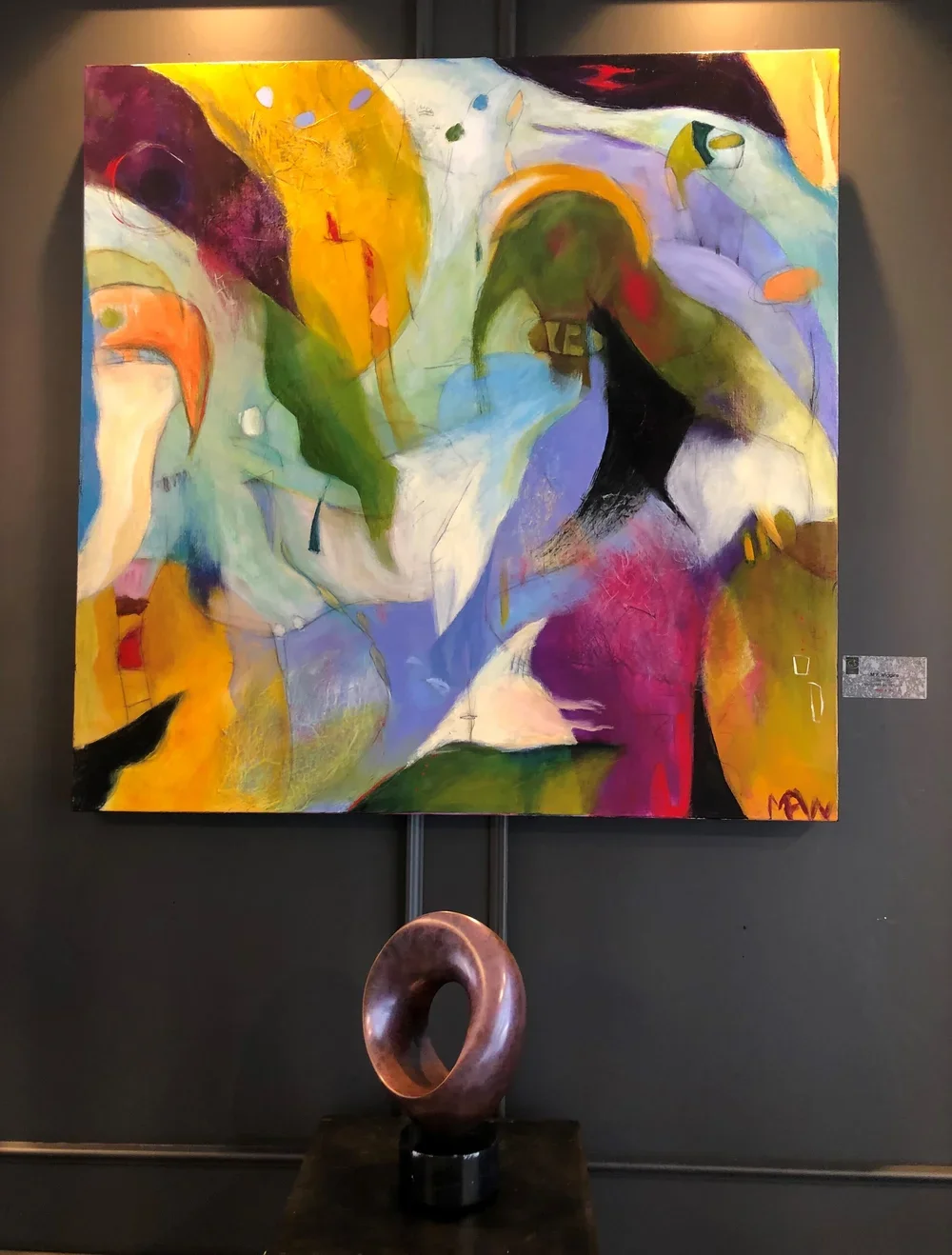 An abstract painting with vibrant colors including yellow, purple, green, black, red, and white, featuring various shapes and brushstrokes. A bronze sculpture of a donut-shaped object is displayed below the painting.