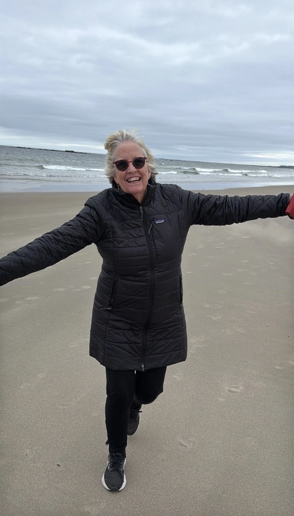 Woman in black jacket and sunglasses running on a beach with arms outstretched, ocean waves in the background, overcast sky.