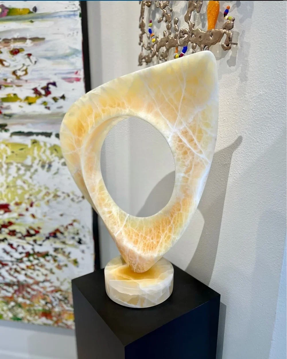 A yellow marble sculpture with a circular hollow center, mounted on a black pedestal against a white wall with abstract art and decorative wall pieces.