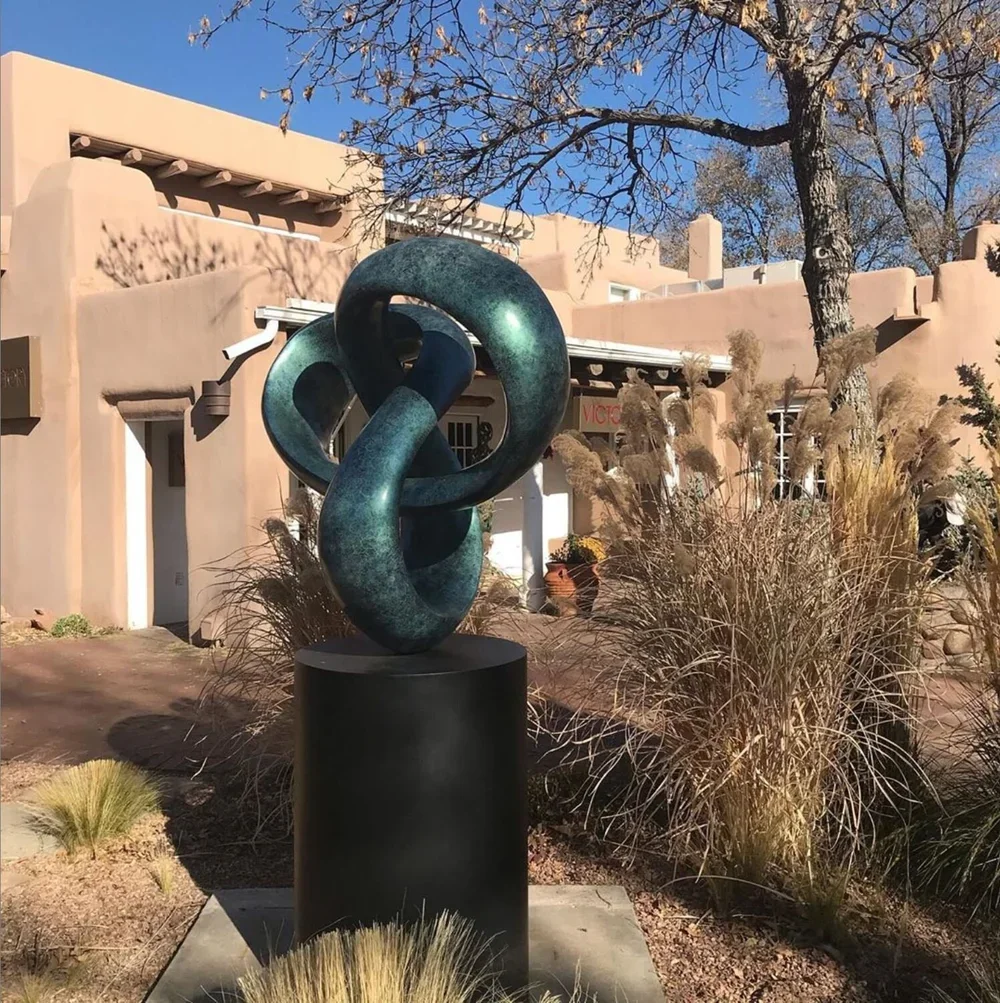 Abstract metal sculpture with twists and loops, situated outdoors in front of a beige stucco building, surrounded by desert plants, dry grasses, and trees under a clear blue sky.