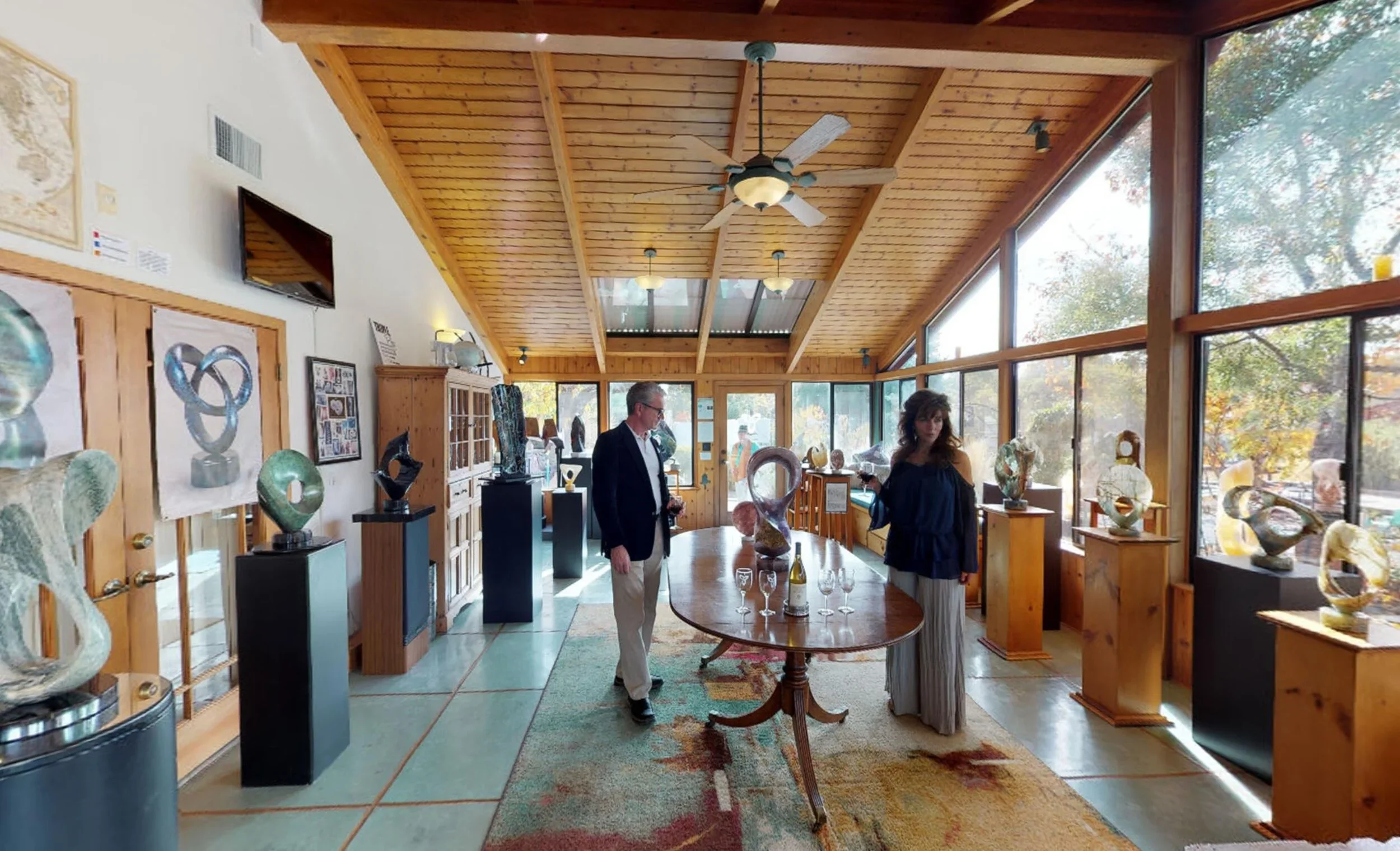 People in an art gallery viewing various abstract sculptures. The gallery has wooden walls, a vaulted wooden ceiling with a ceiling fan, large windows on one side allowing natural light, and display pedestals and cabinets with art pieces.