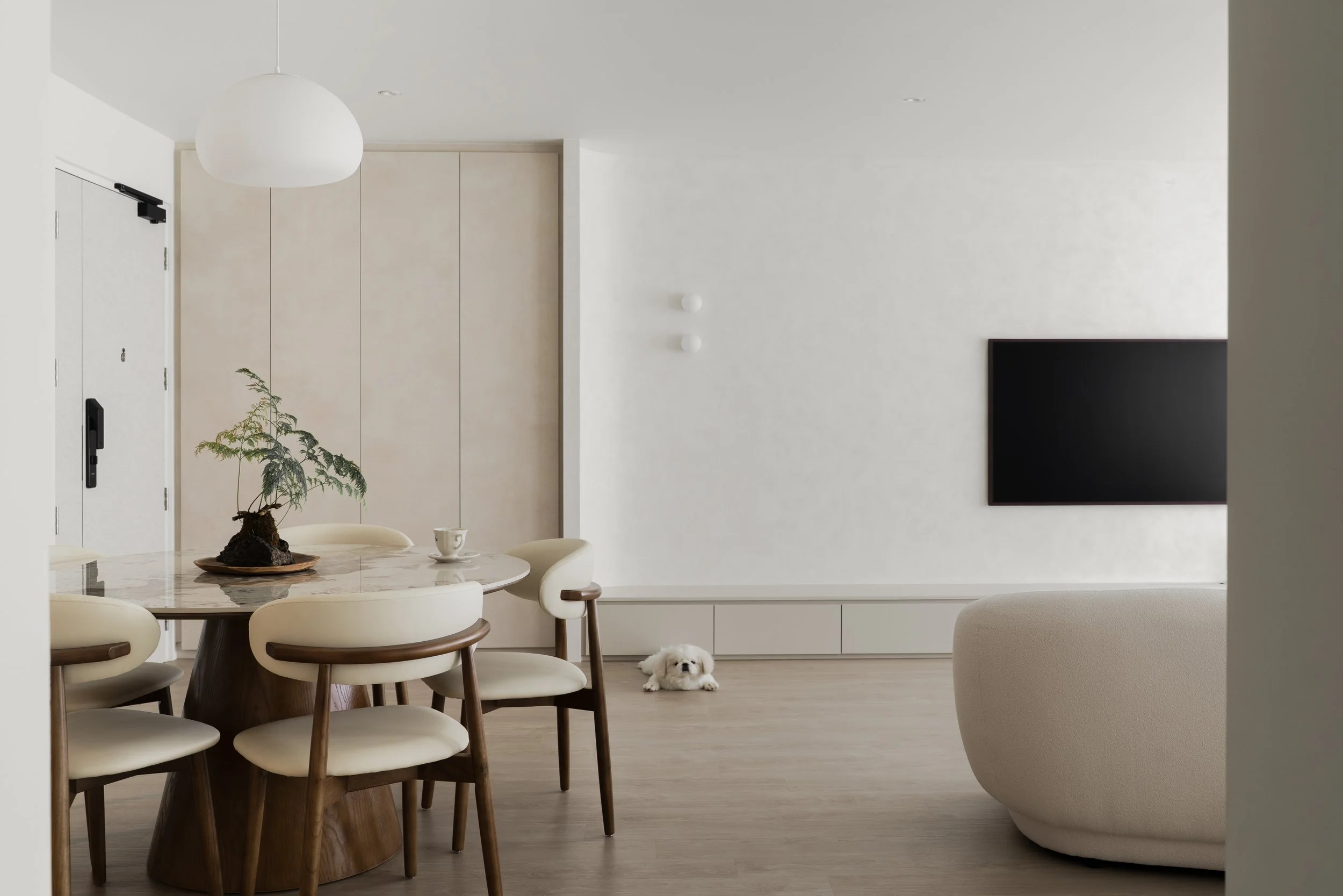 Minimalist 5-room BTO Tampines living room with a dining table, a potted plant, a small white dog on the floor, a large flat-screen TV on the wall, and neutral-colored furniture, designed by Built Different, an interior design firm in Singapore.