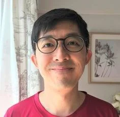 A person wearing a dark jacket and khaki pants standing in tall yellow grass, looking towards distant mountains under a cloudy sky.