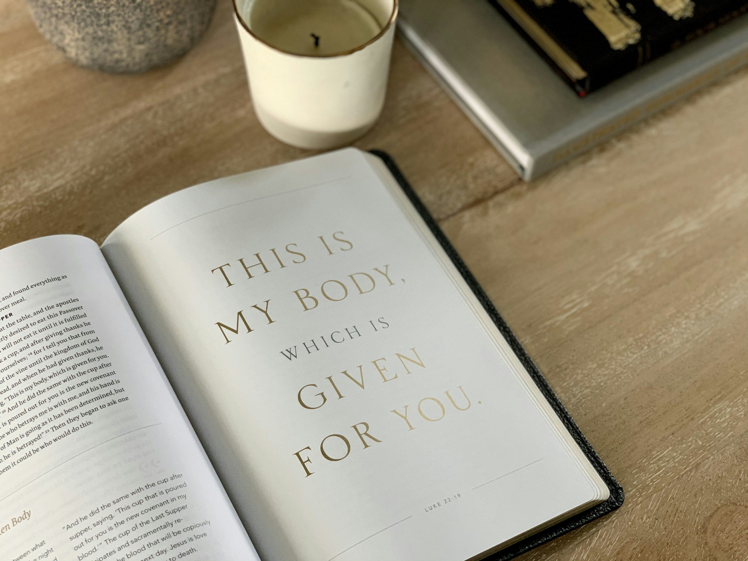 Open Bible with a passage that says 'This is my body, which is given for you' on the right page, placed on a wooden table with a candle, a stone object, and a stack of books nearby.