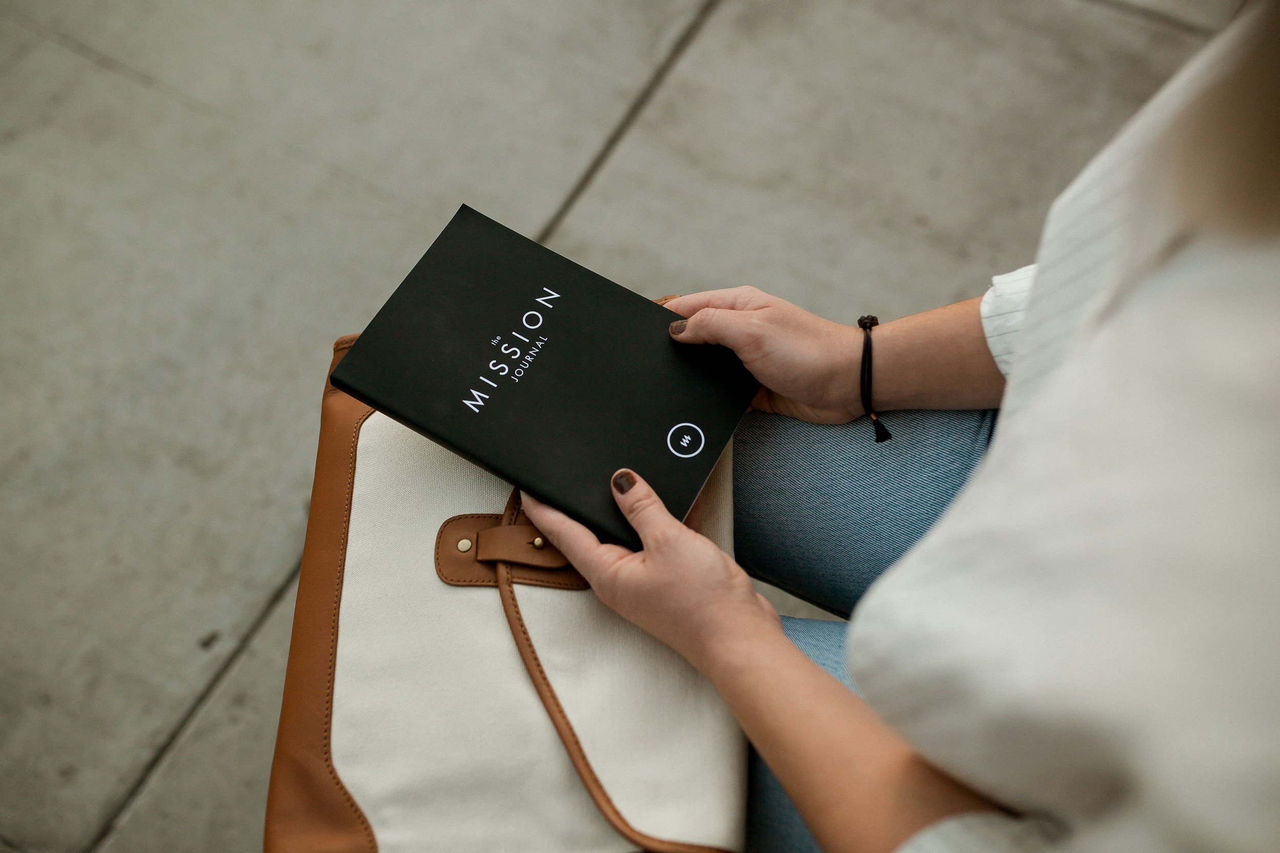 A woman sitting on a beige and brown chair, holding a black journal titled 'MISSION Journal.' She has a black bracelet on her wrist and is wearing a white shirt and blue jeans.