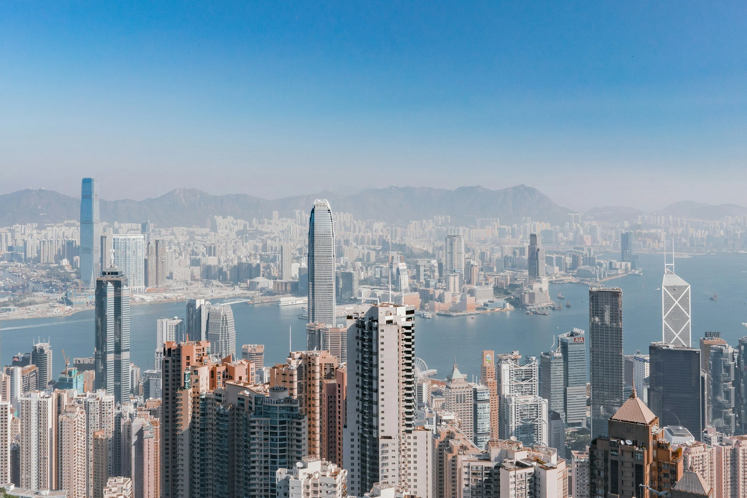 Aerial view of Hong Kong city skyline with tall skyscrapers and Victoria Harbour, mountains in the background under a blue sky.