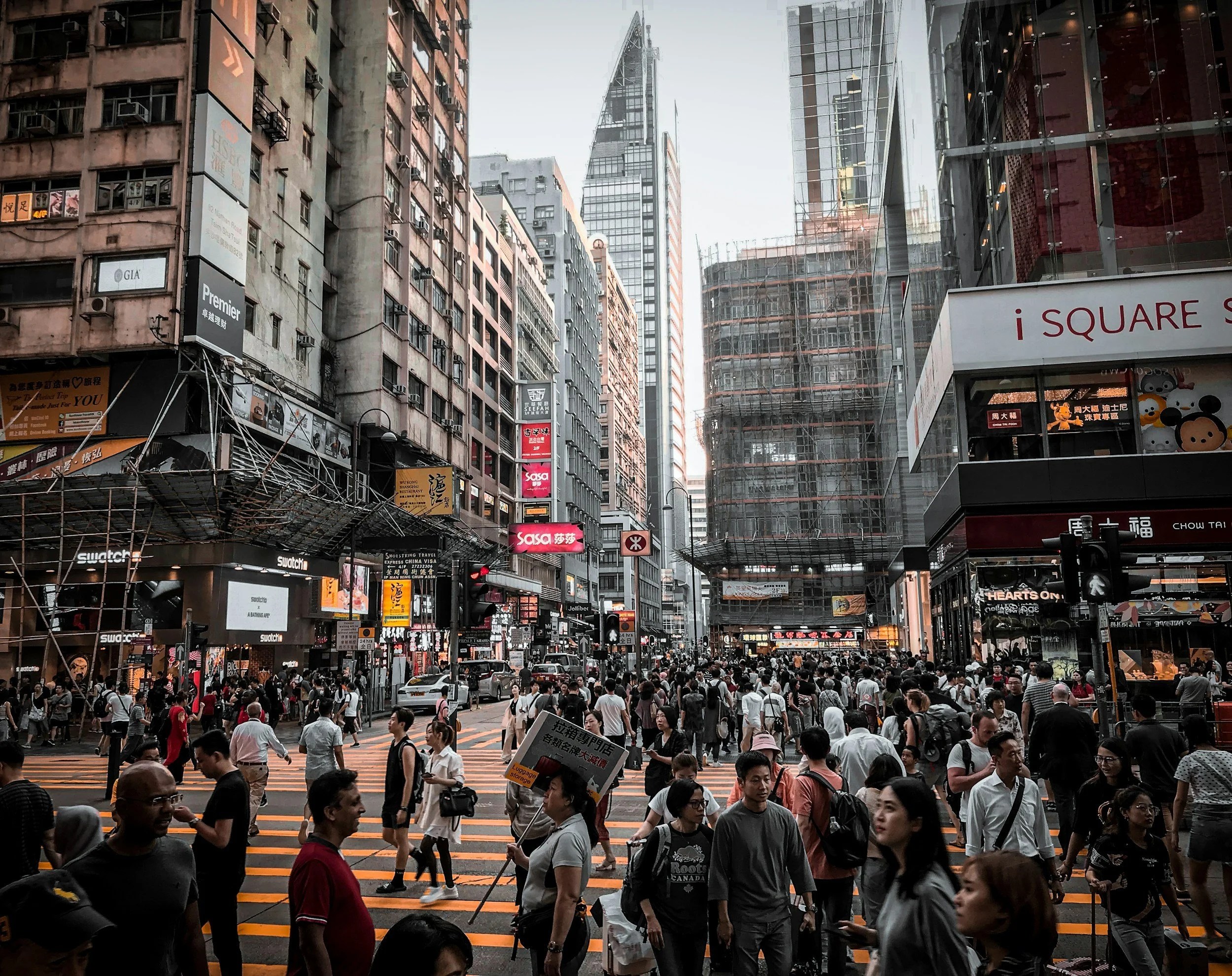 A busy urban street scene in Hong Kong with many pedestrians crossing the street at a crosswalk, surrounded by tall buildings with illuminated signs and advertisements.