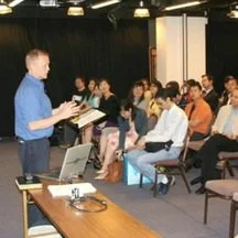 A man in a blue shirt giving a presentation to a seated audience in a conference room, glimpse of ECC HK