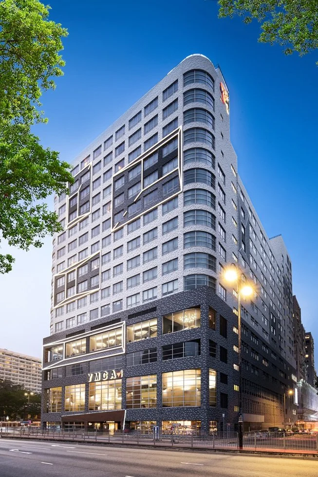 Modern multi-story building with glass windows, geometric design accents, and streetlights in an urban area at dusk. YMCA, Tsim Sha Tsui, Hong Kong