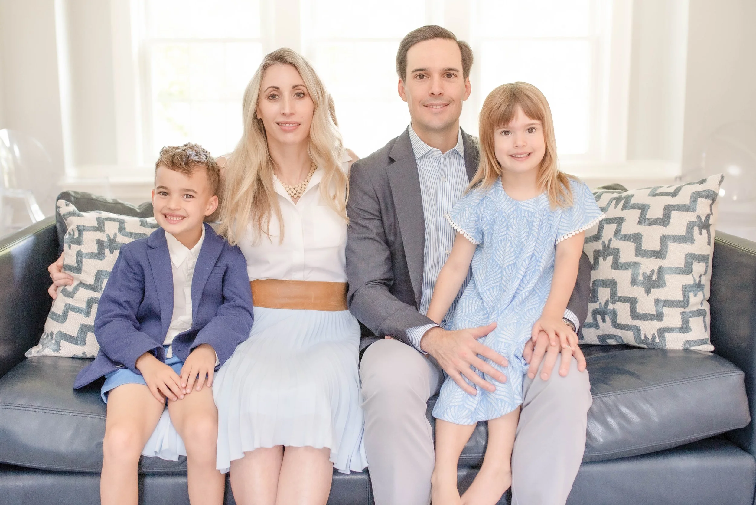 A family of five sitting on a sofa in a bright room. Two children, a boy and a girl, sit with their parents, smiling at the camera.