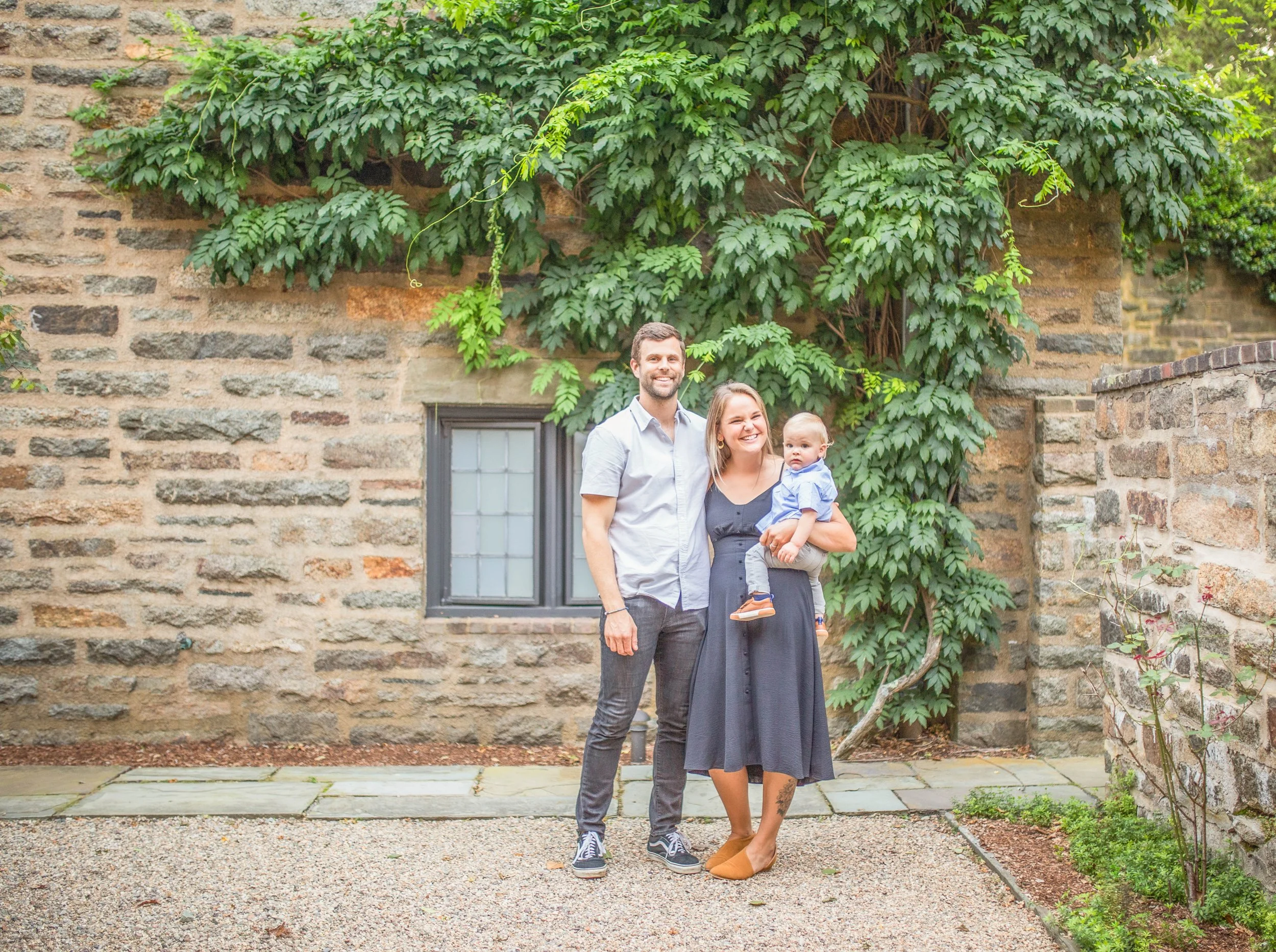 A family of three standing outdoors in front of a stone and brick wall with green ivy and plants. The family includes a man, a woman holding a young child, all smiling.