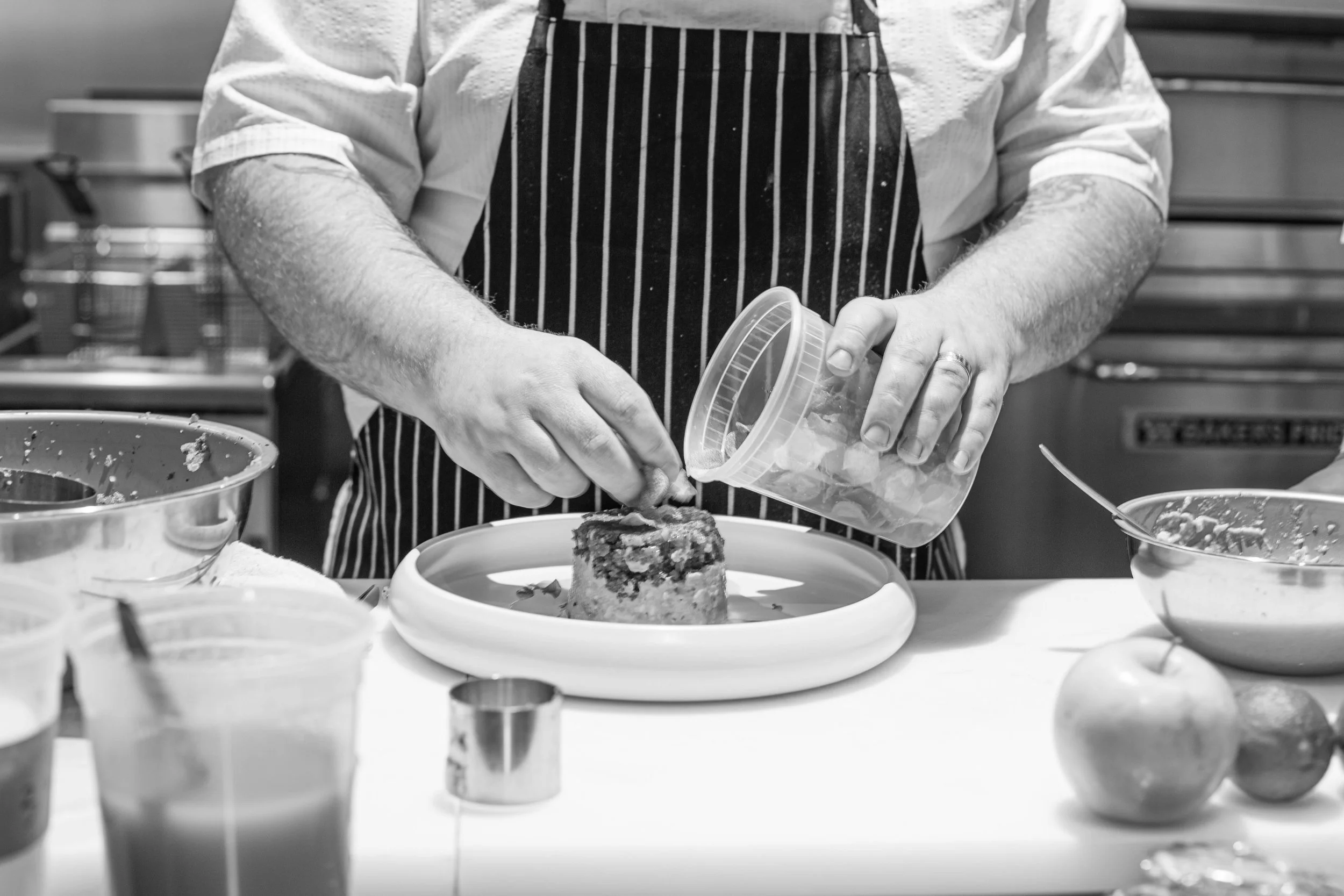 A chef in a striped apron decorates a cake with toppings while icing a cake on a round plate.