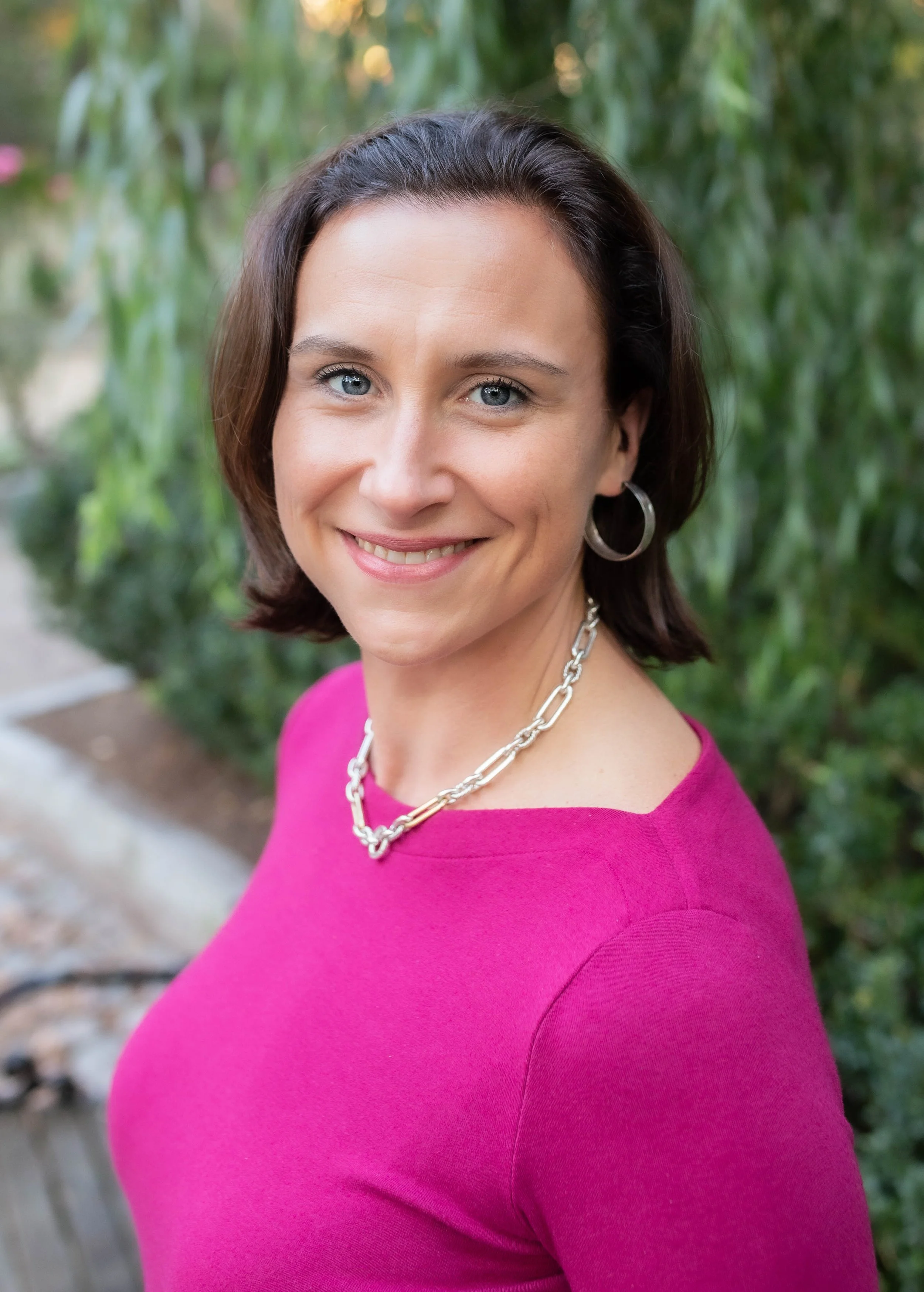 A smiling woman with short dark hair, wearing a pink top, silver chain necklace, and hoop earrings, standing outdoors with greenery in the background.