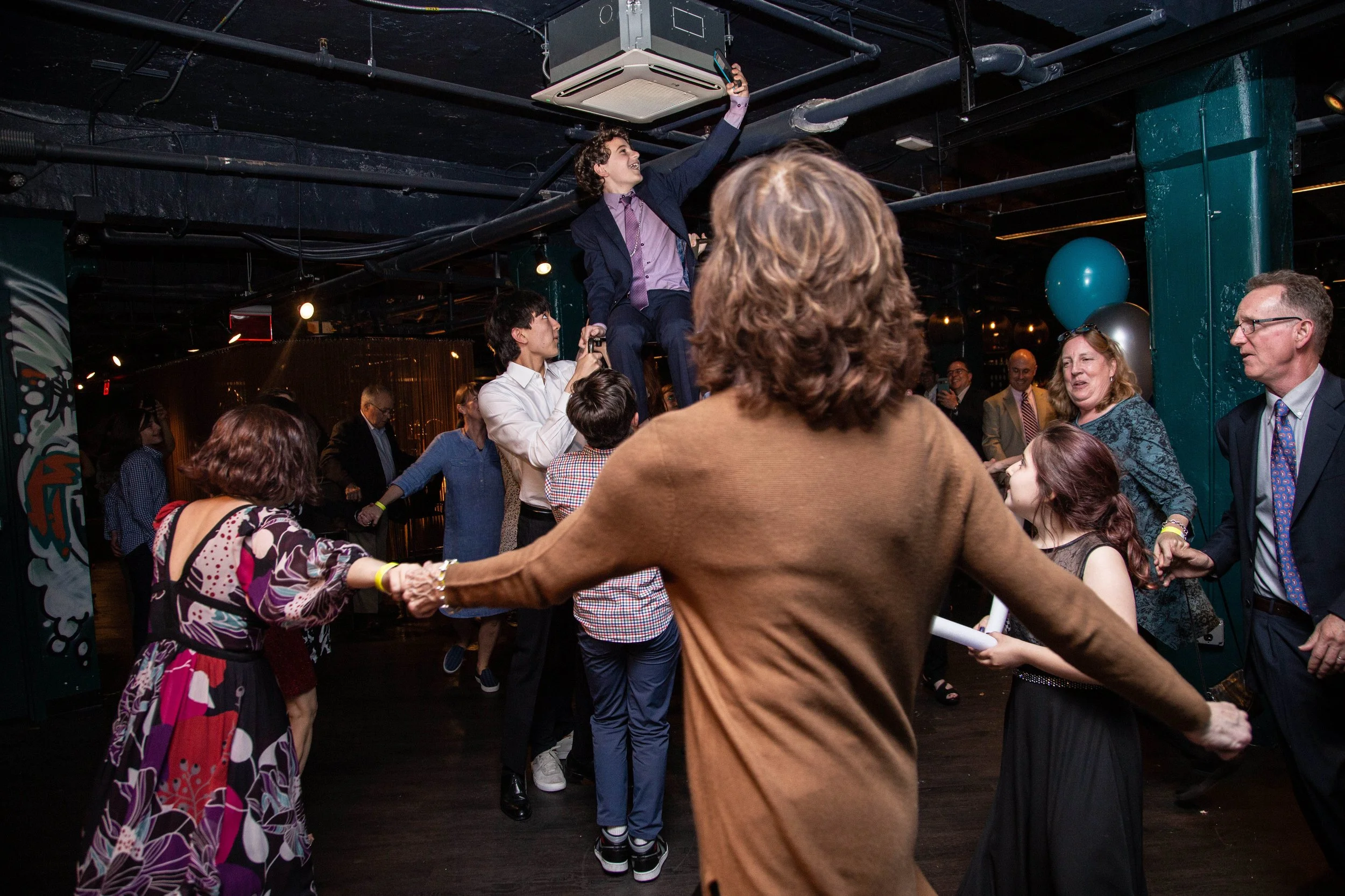 People dancing and holding hands at a party with a person sitting on a chair being lifted above the crowd.