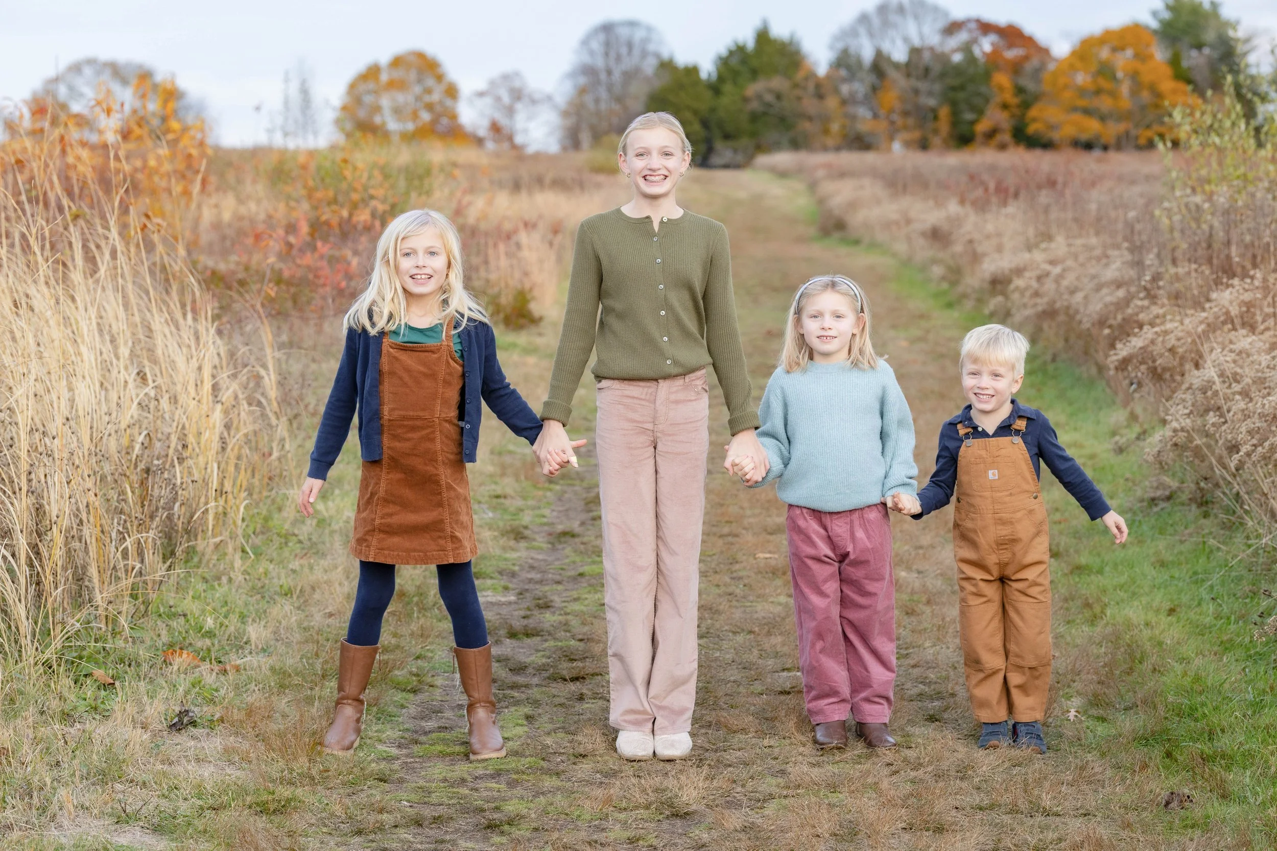 Four children, two girls and a boy, holding hands and walking on a dirt path in an autumn landscape with colorful trees in the background.