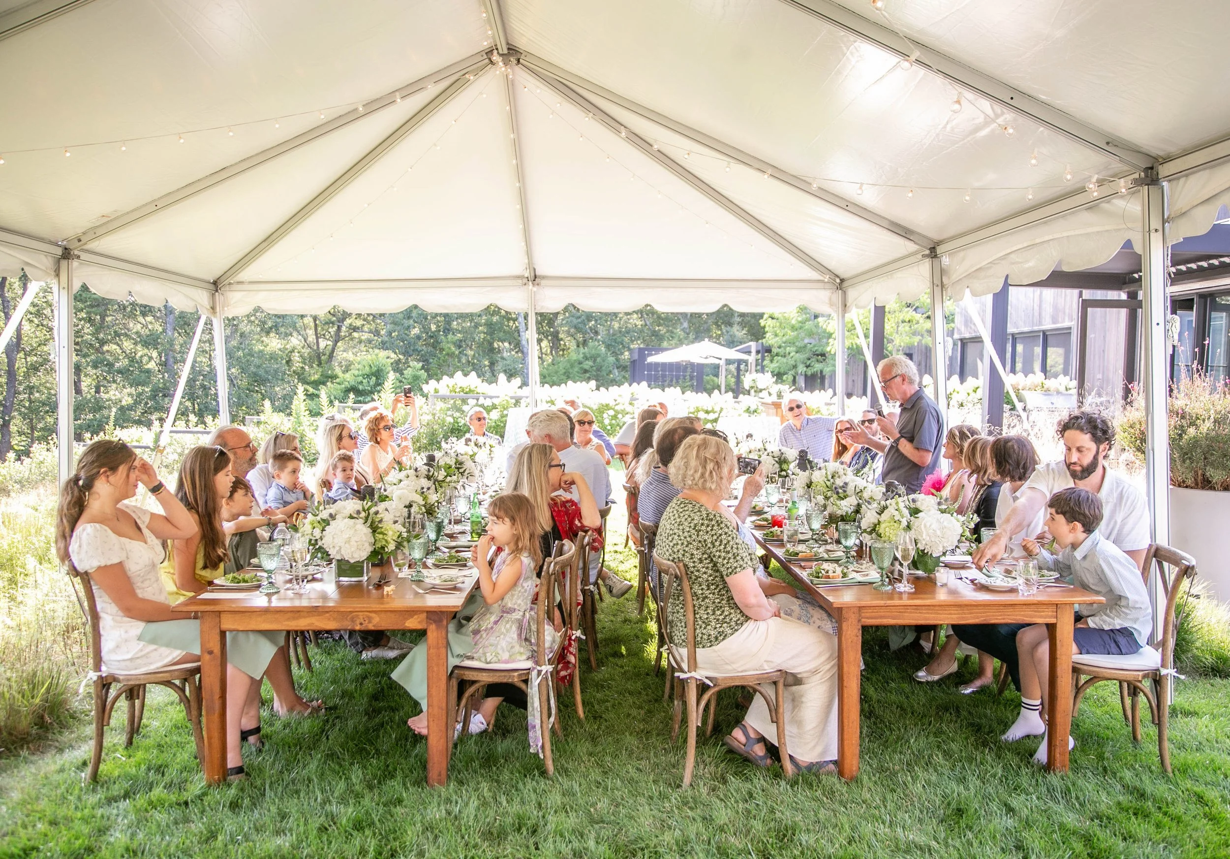 People gathered around two long tables enjoying a outdoor celebration under a white canopy tent with floral centerpieces.