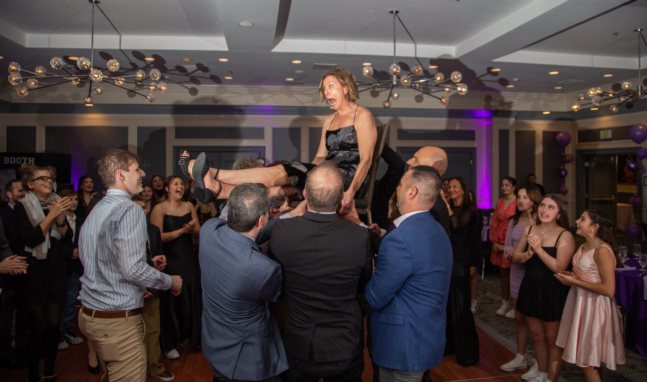 Group of people celebrating at a party, lifting a woman in the air and everyone cheering and clapping, with purple lighting and festive decorations in the background.
