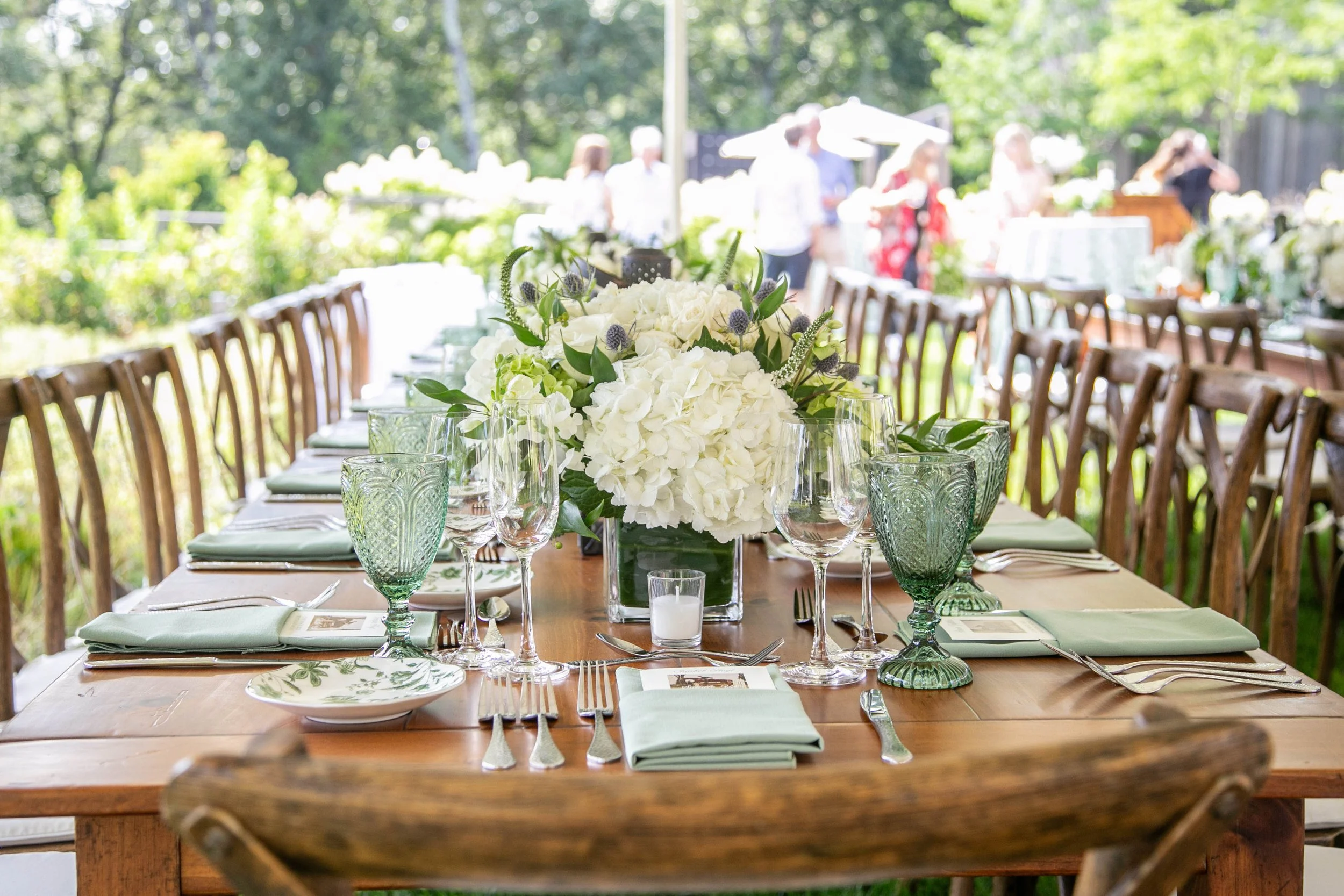 Outdoor dining table decorated with white flowers, green glassware, and silverware, set for a meal on a sunny day. Blurred guests are in the background.