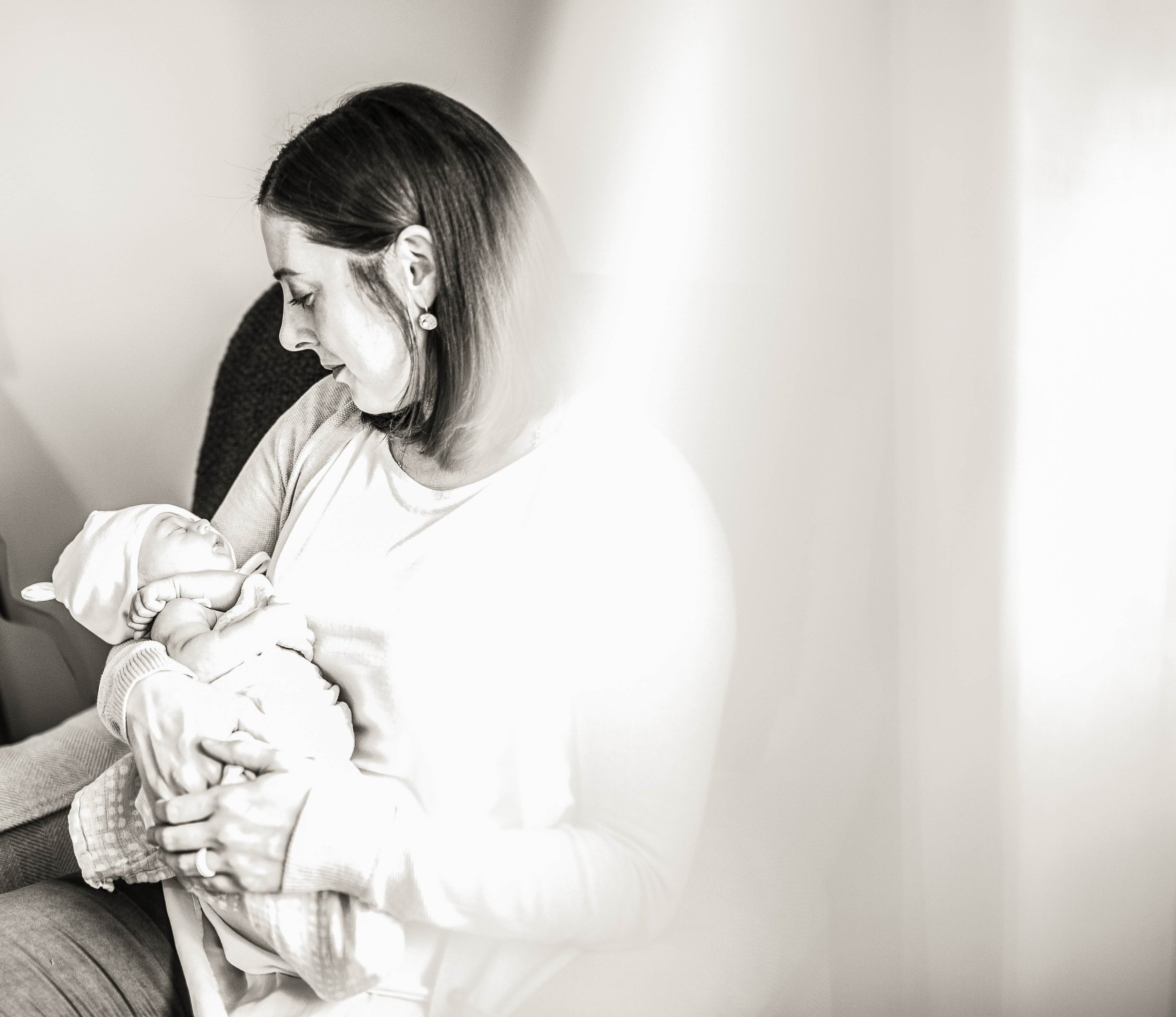 A woman holding a newborn baby in her arms, sitting on a chair, looking down at the baby lovingly.