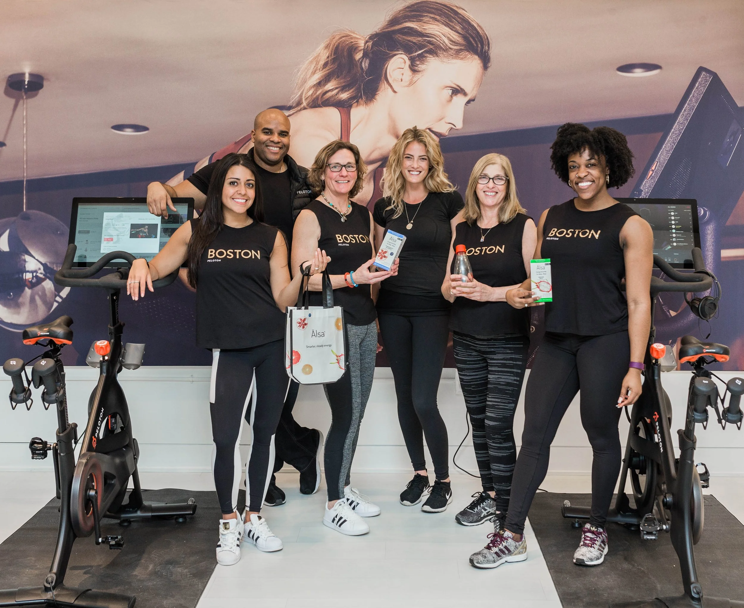 Group of six smiling women and one man in a fitness studio, holding fitness products, with exercise bikes and a large poster of a woman on the wall behind them.