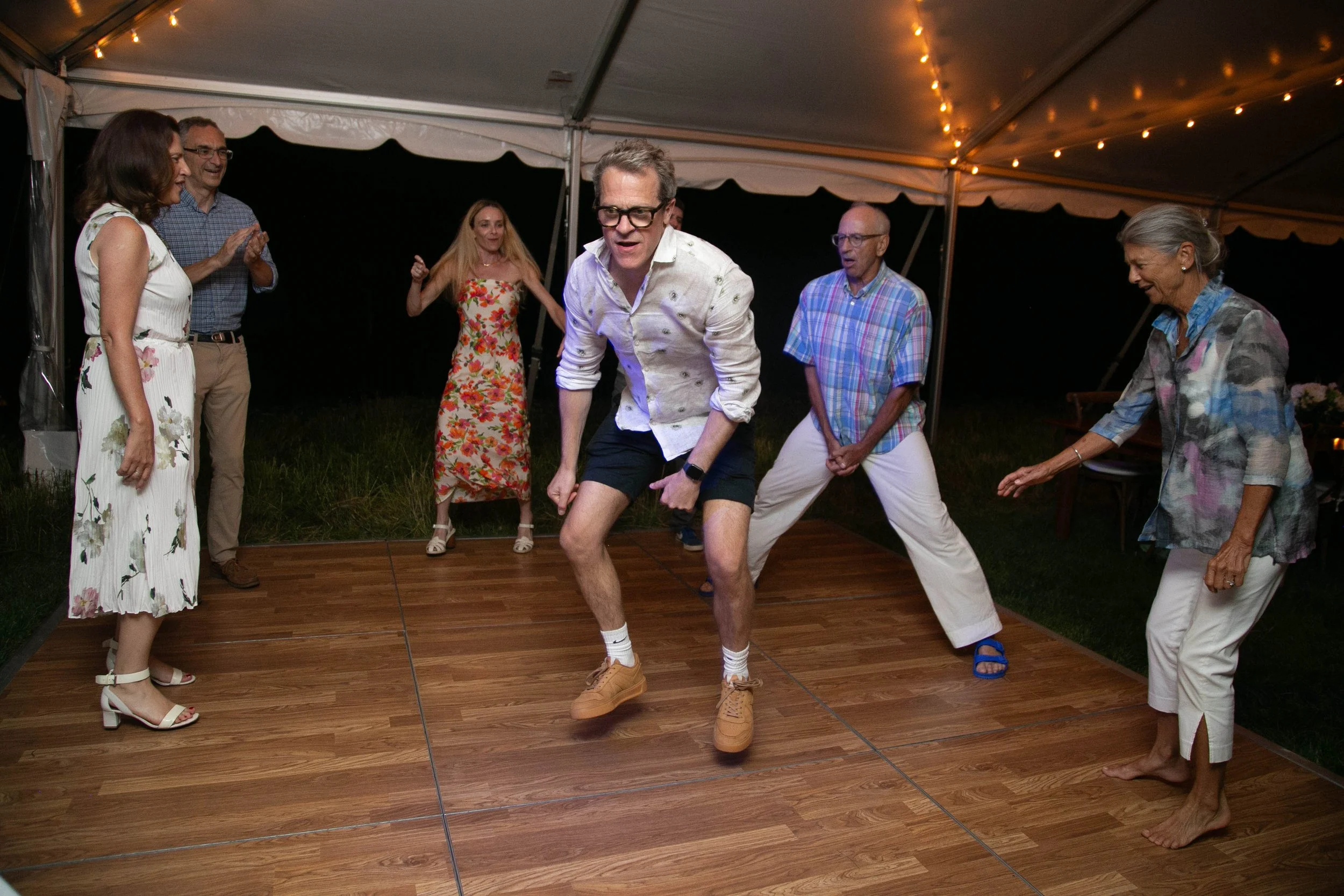Group of people dancing on a wooden floor under a tent at night, with string lights overhead, celebrating and enjoying themselves.