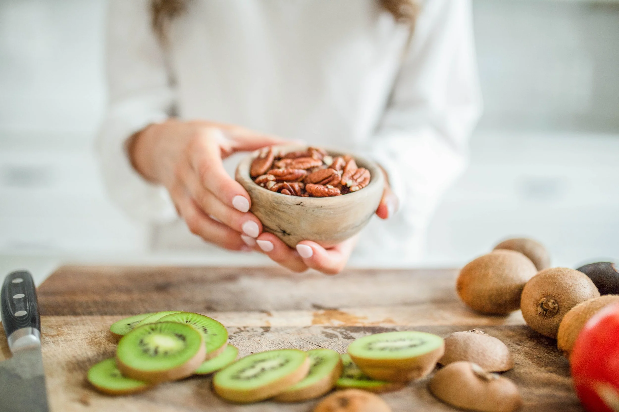 Person holding a wooden bowl filled with pecans over a chopping board with sliced kiwi, whole kiwis, and a knife, in a bright kitchen setting.