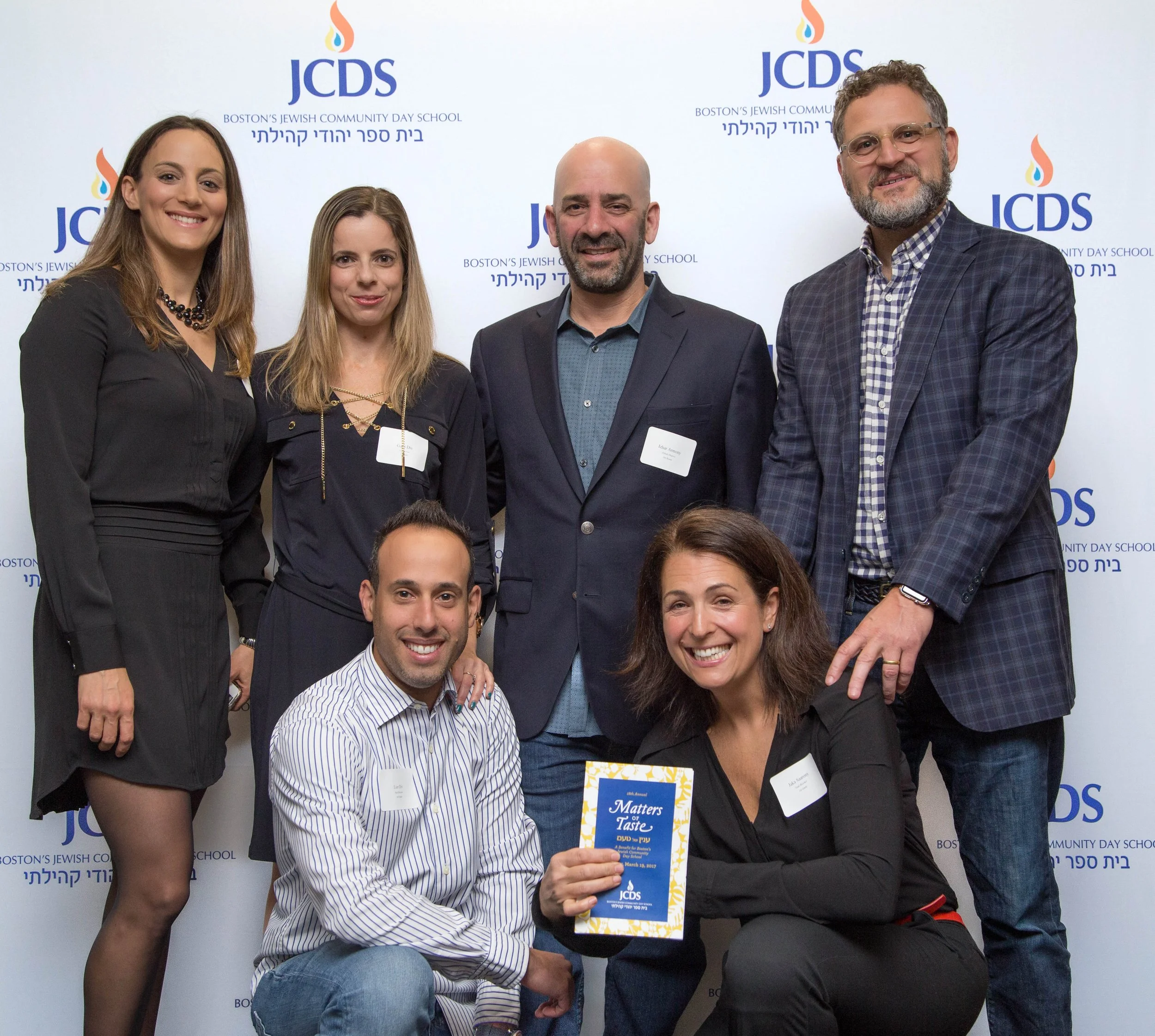 A group of seven people, including men and women, posing together at an event, with a backdrop displaying the logo and text for JCDS and Boston's Jewish Community Day School.