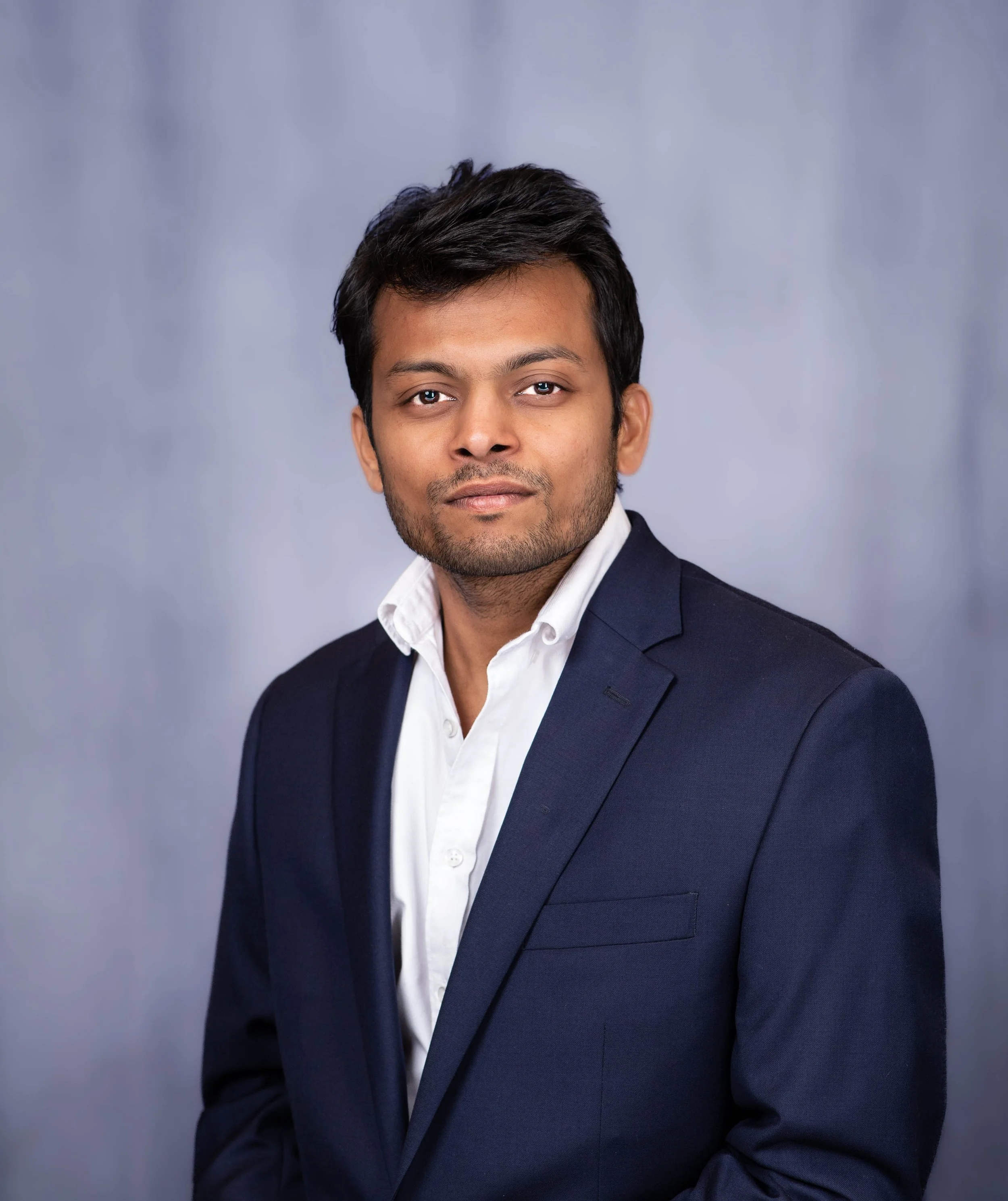 A professional young man in a dark suit and white shirt posing against a plain gray background.