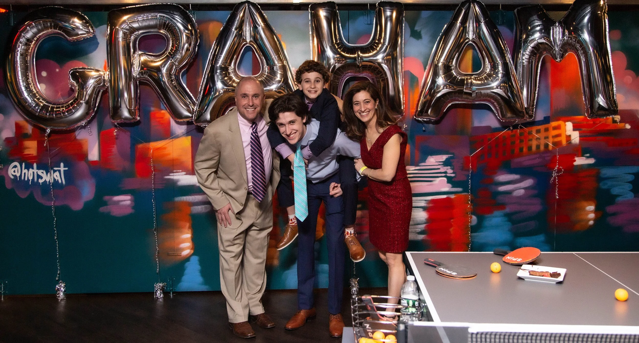A group of four adults and one child smiling and posing together at a celebration, standing in front of balloons spelling 'GRADUATION.' The background has colorful, blurred cityscape artwork, and there is a ping pong table on the right with ping pong