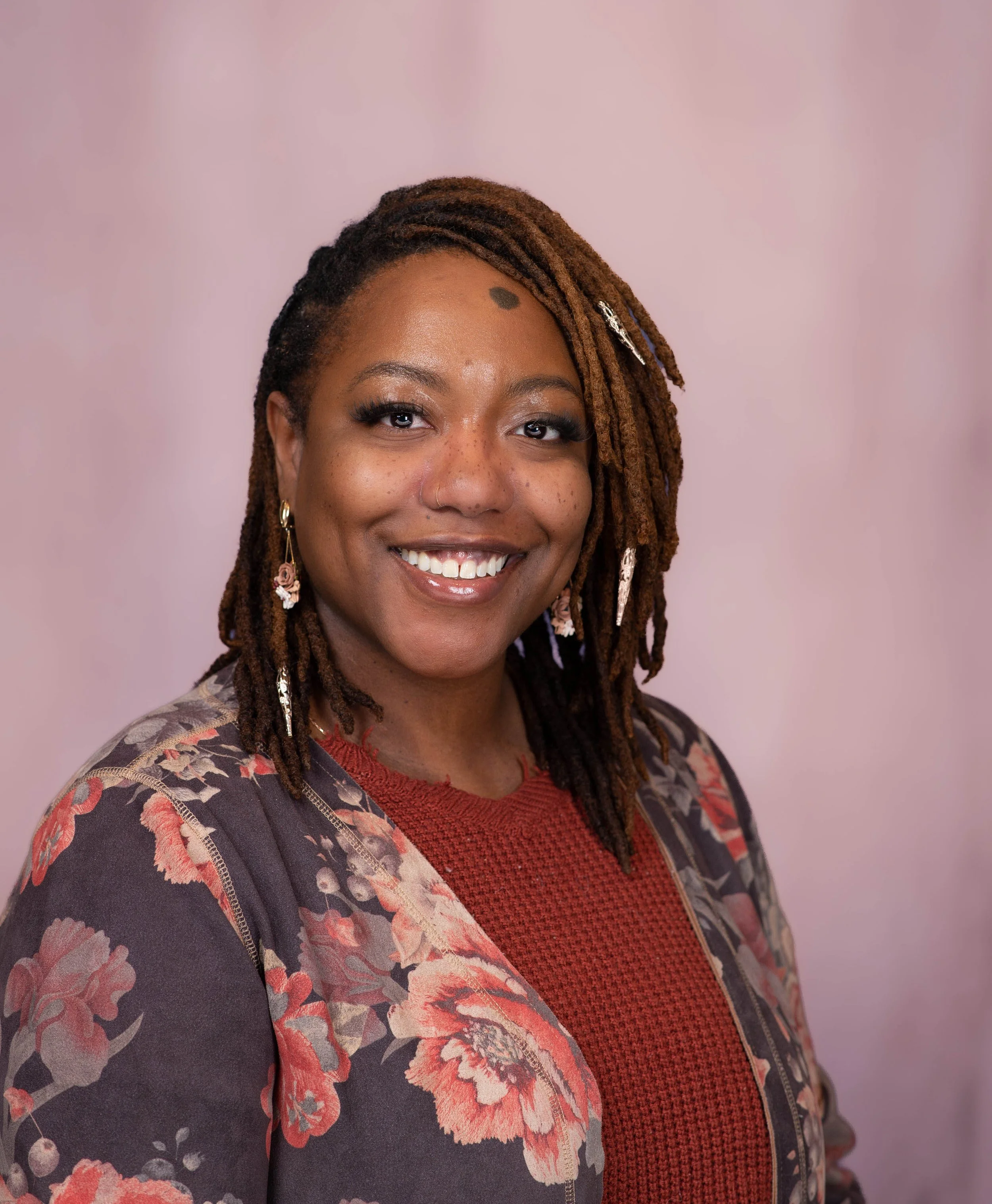 A woman with brown dreadlocks, earrings, and a floral blazer, smiling against a pink background.
