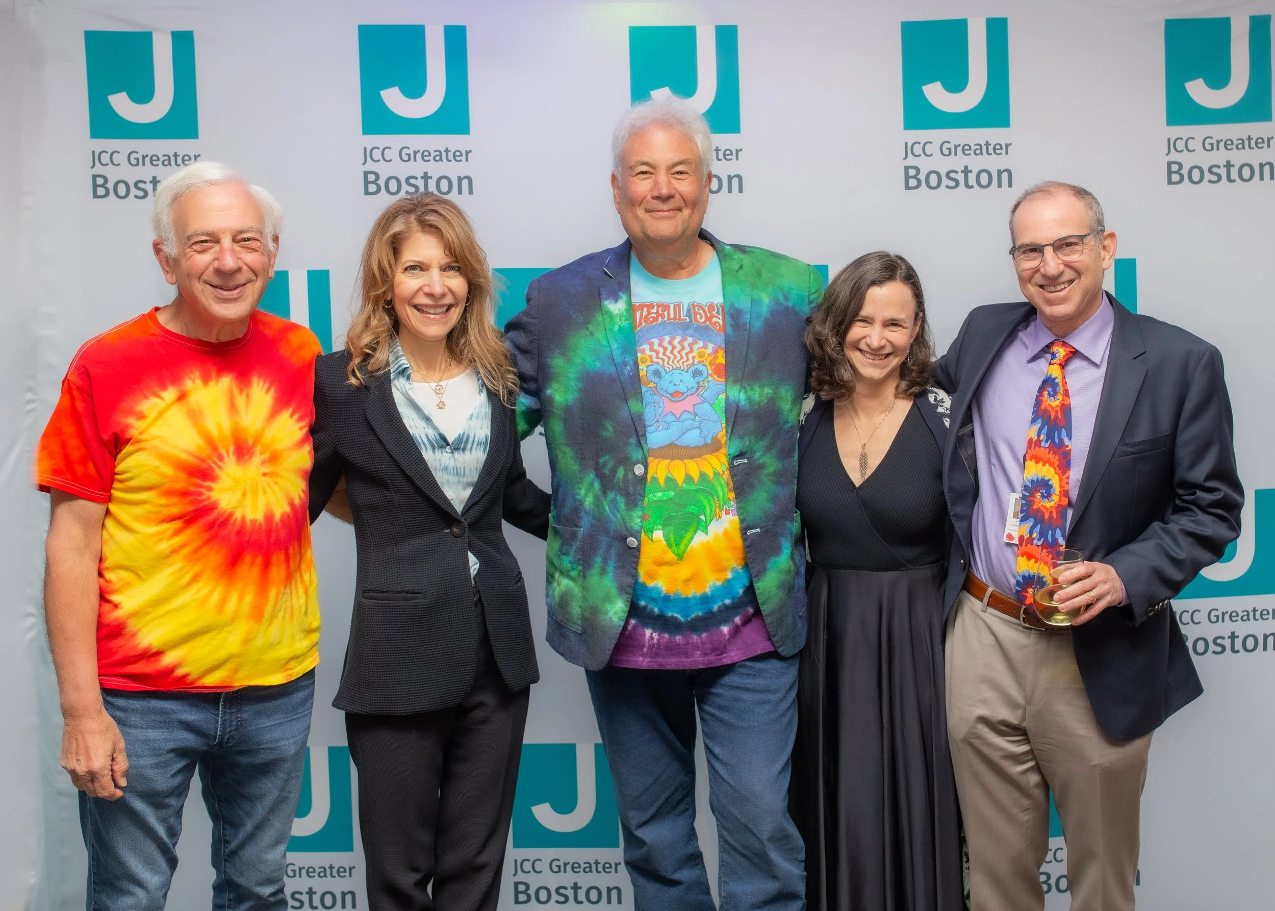 Group of five people standing together in front of a backdrop with the JCC Greater Boston logo. They are smiling and dressed in colorful, casual, and semi-formal attire, with some wearing tie-dye shirts.