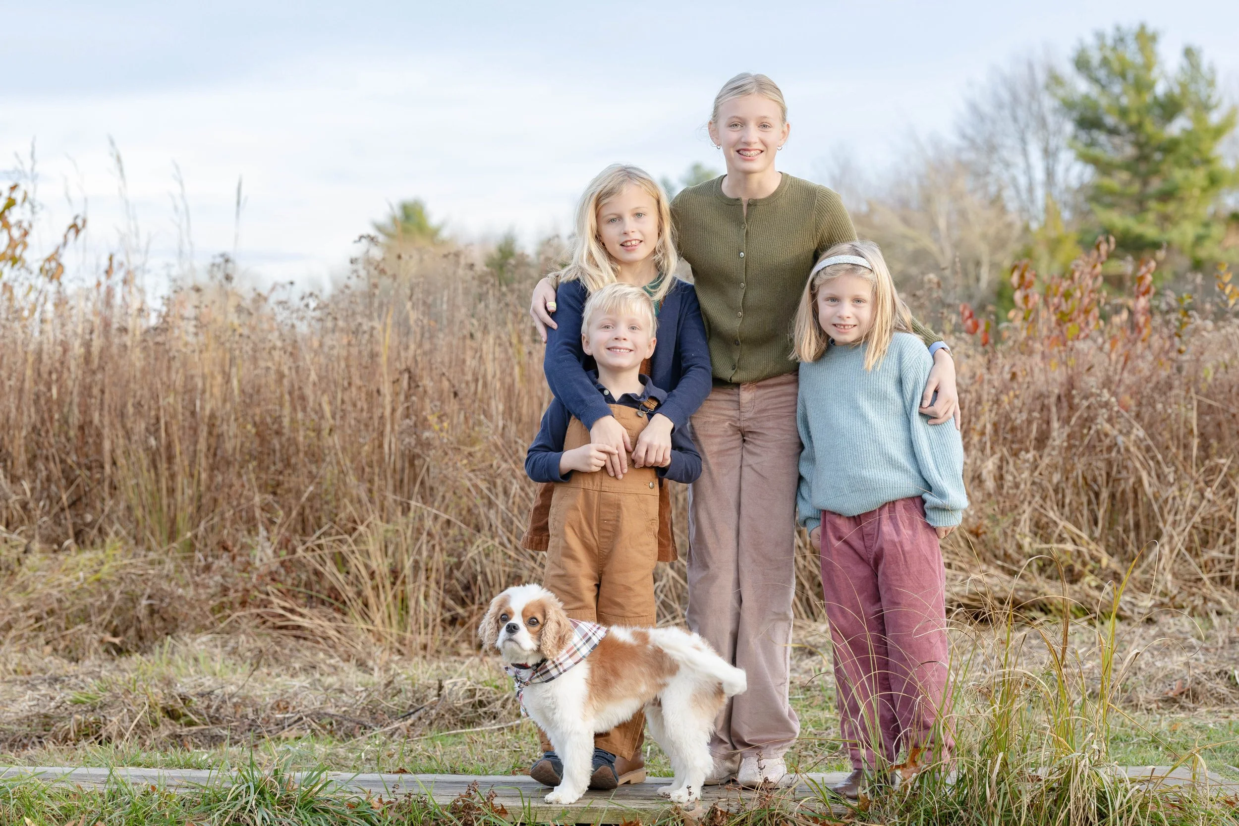 A woman with three children and a dog standing outdoors in a field with tall dry grass and trees in the background.
