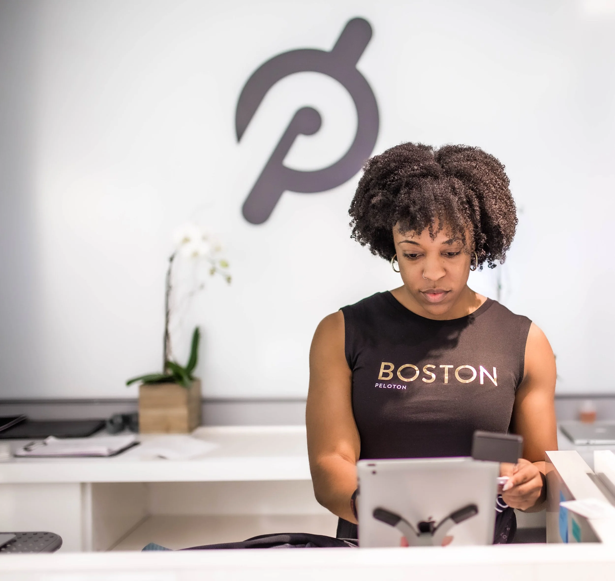 A woman with curly hair and ear piercings using a tablet at a Peloton fitness studio, with a Peloton logo on the wall behind her.