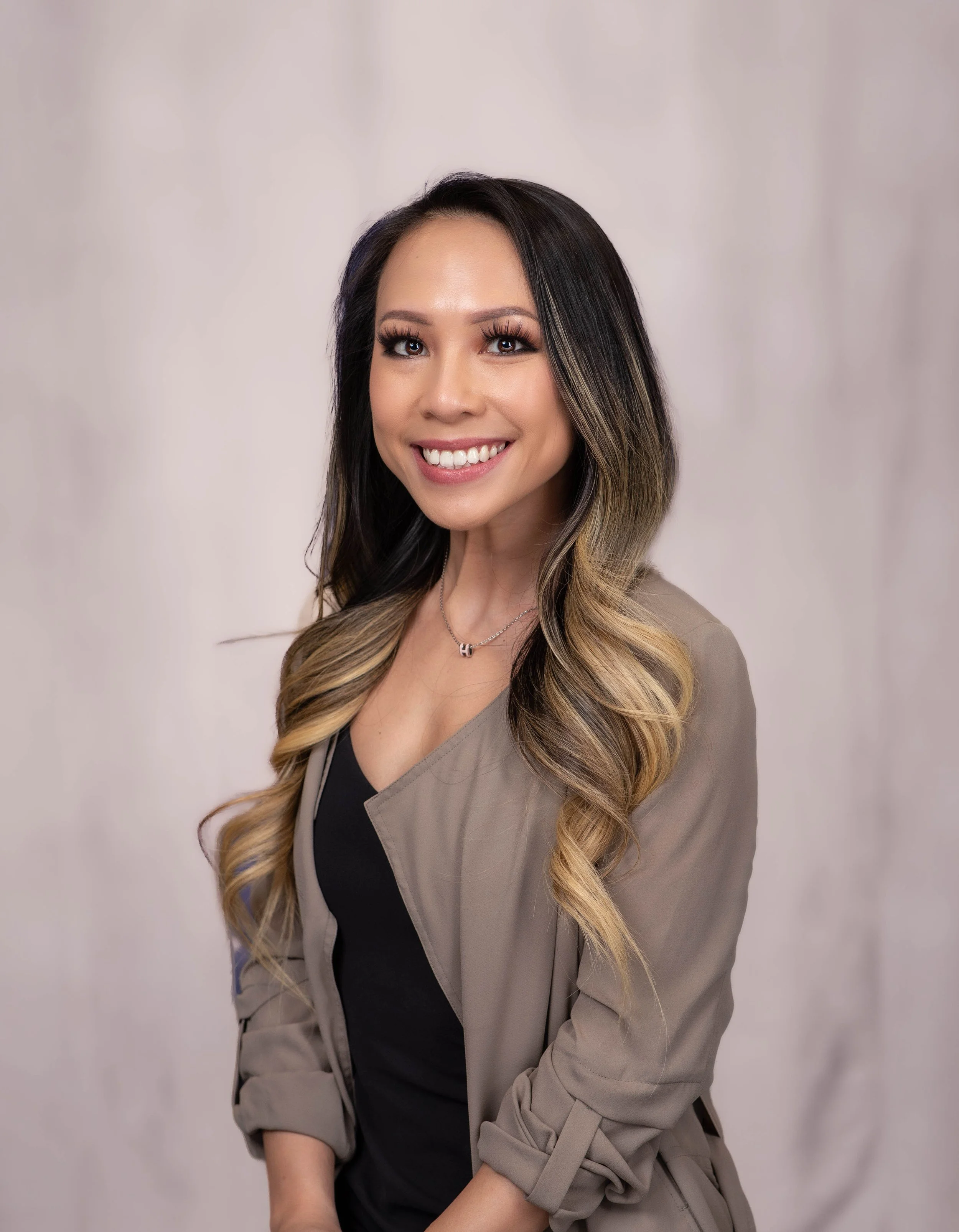 A smiling woman with long wavy hair and a beige blazer over a black top, posing against a neutral background.