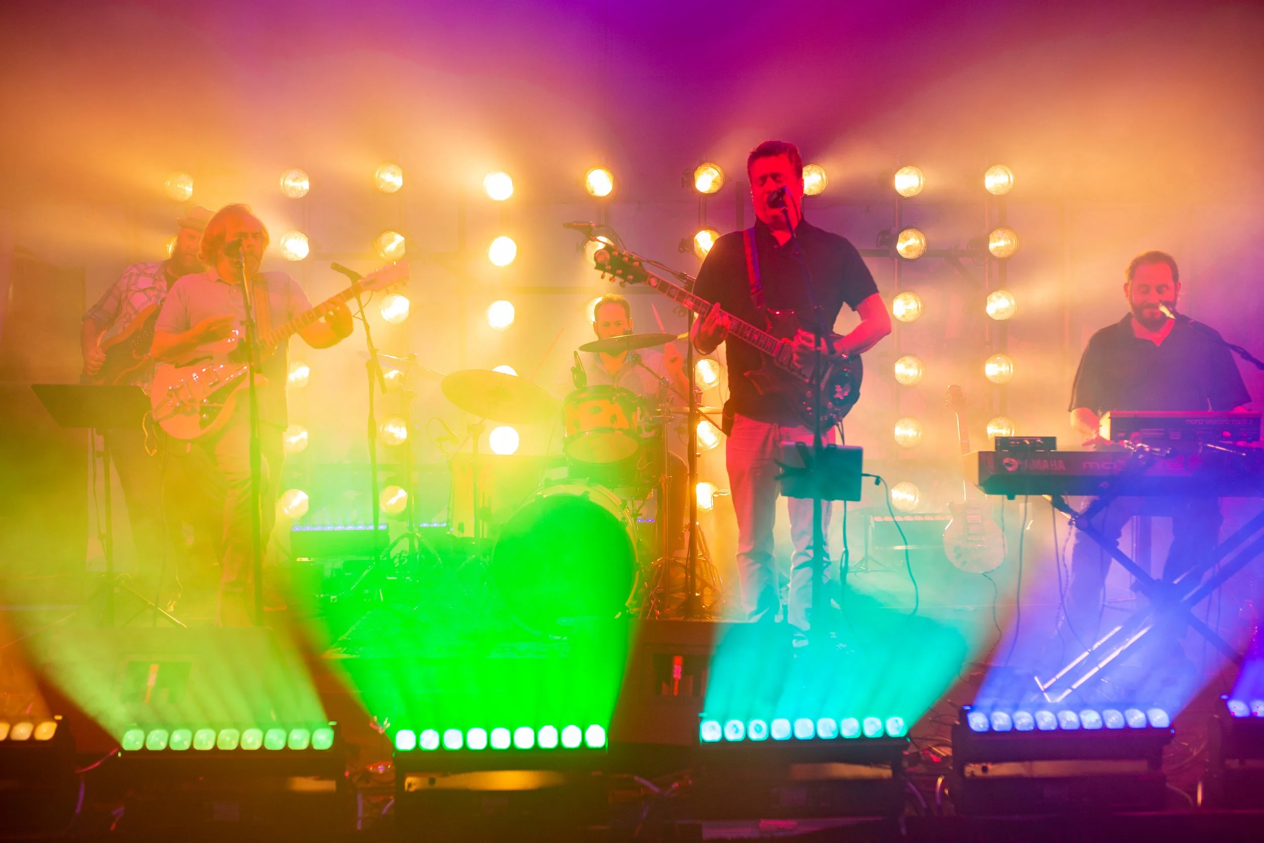 A band performing on stage with colorful stage lights, including two guitarists, a drummer, and a keyboardist.