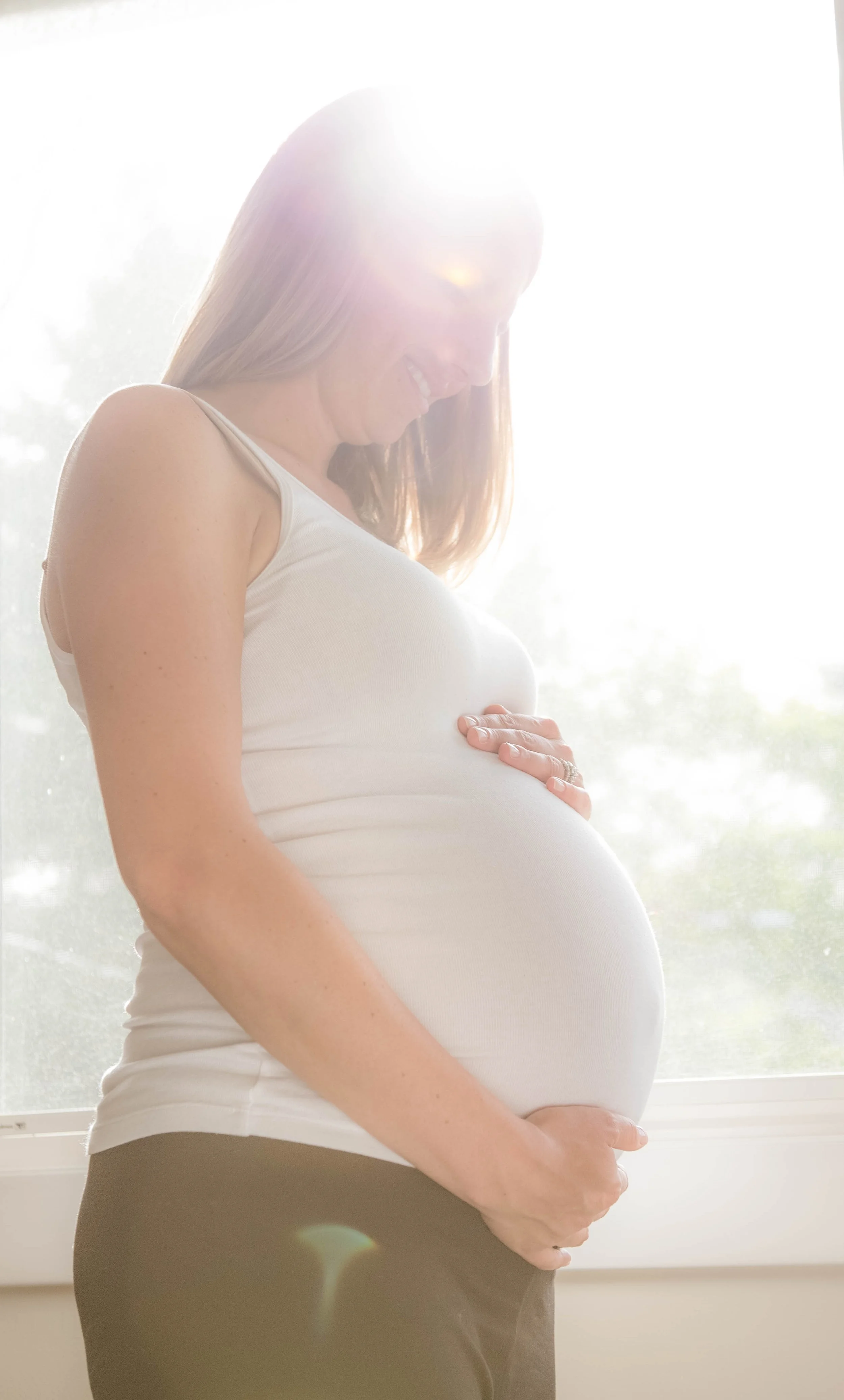 A pregnant woman standing in front of a bright window, smiling and gently holding her baby bump.