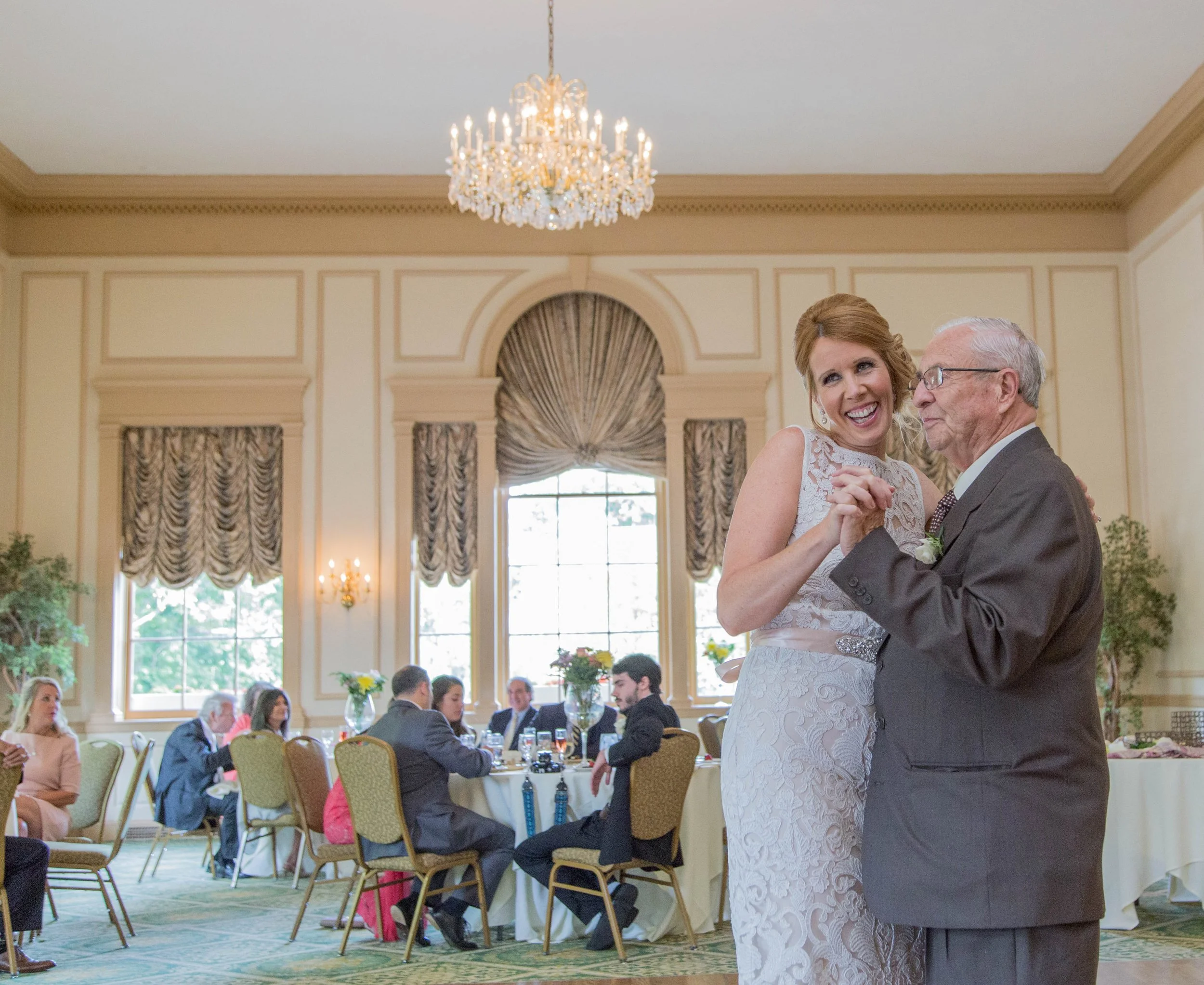 A bride dancing with an elderly man at a wedding reception in a grand hall with large windows, decorated with chandeliers and floral arrangements.